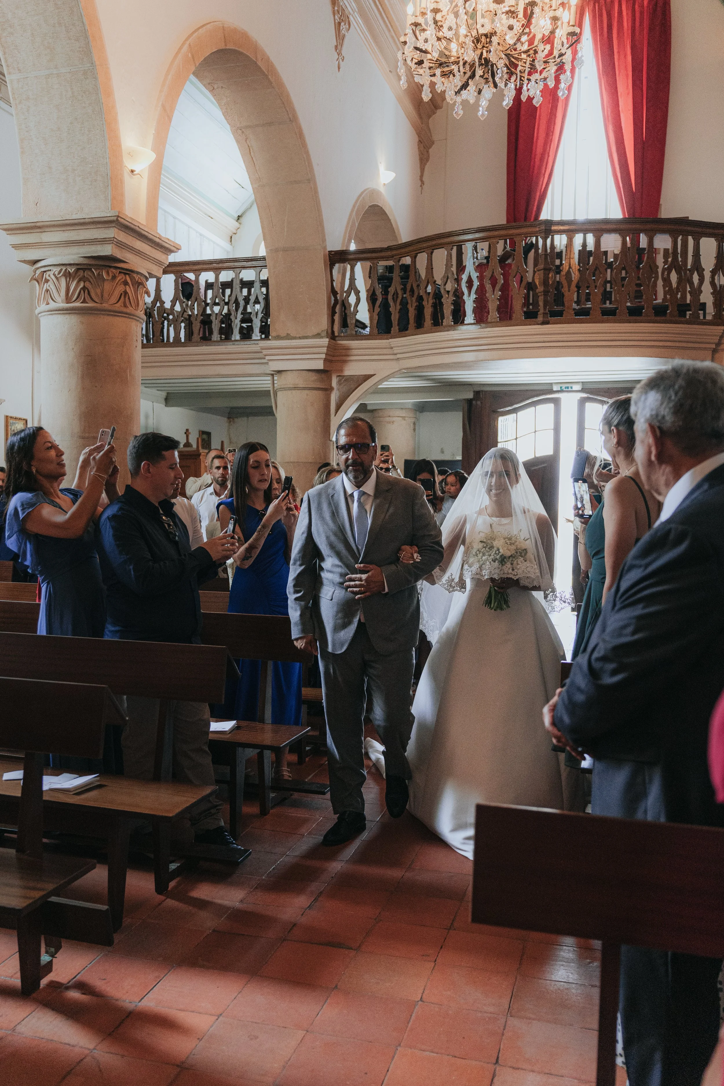 A wedding ceremony inside a church with guests standing and taking photos. The bride is wearing a white dress and veil, holding a bouquet, and walking down the aisle. The groom is dressed in a dark suit, standing at the front. The church has high cei