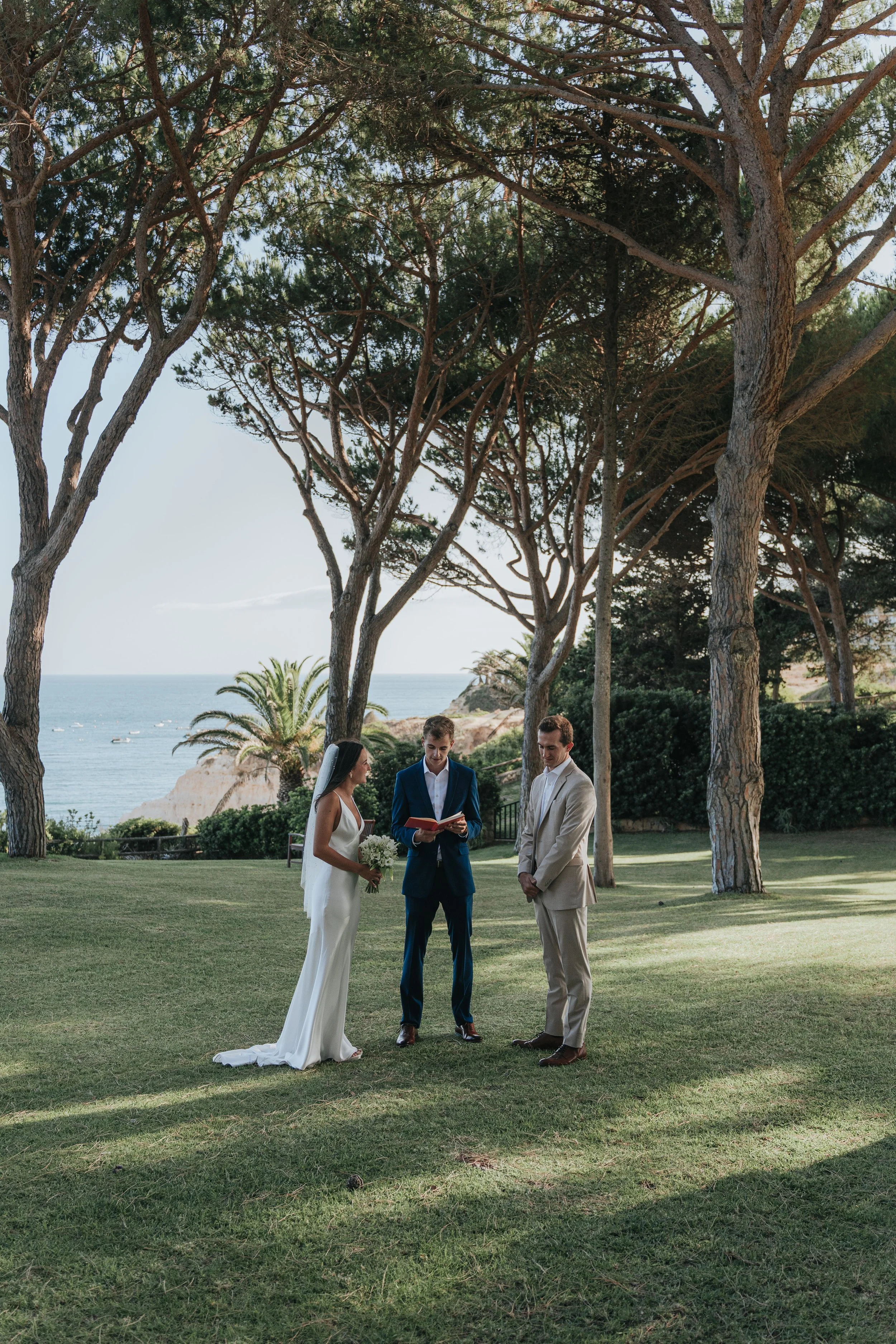 A wedding ceremony taking place outdoors on a grassy area with tall trees, overlooking the ocean. A bride and groom are standing facing each other, with an officiant reading from a book, while the bride holds a bouquet of white flowers.