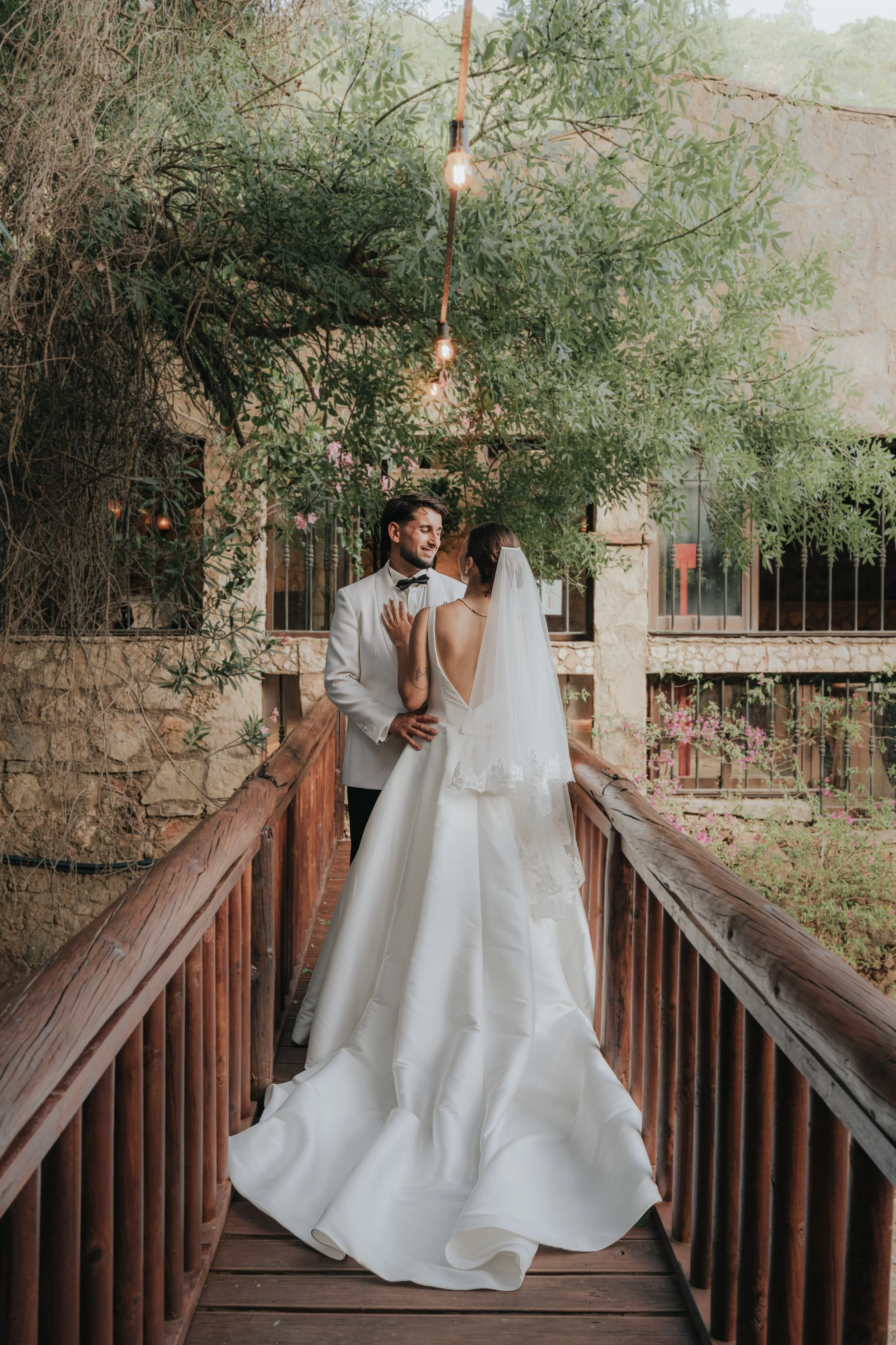 A bride and groom standing on a wooden bridge outdoors, embracing and looking at each other, with lush greenery and a stone building in the background.