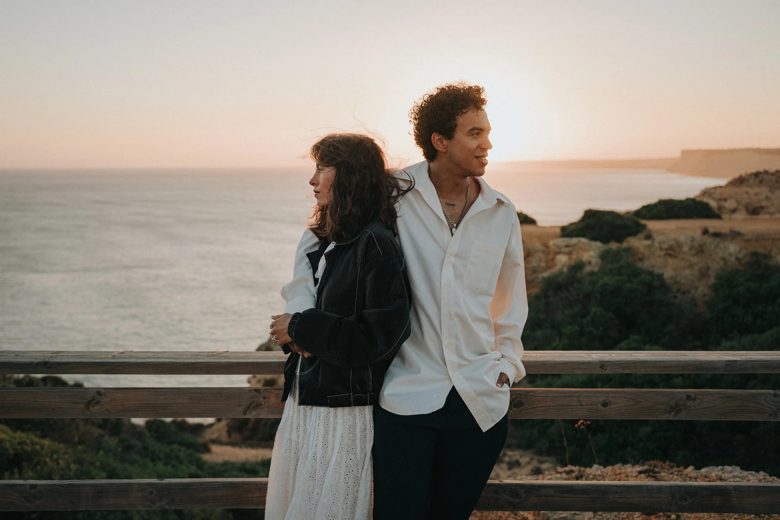 A young man and woman standing near a wooden railing overlooking the ocean at sunset. The woman has dark hair and is wearing a black jacket and light-colored dress, and the man has curly hair and is wearing a white shirt and dark pants. They are look