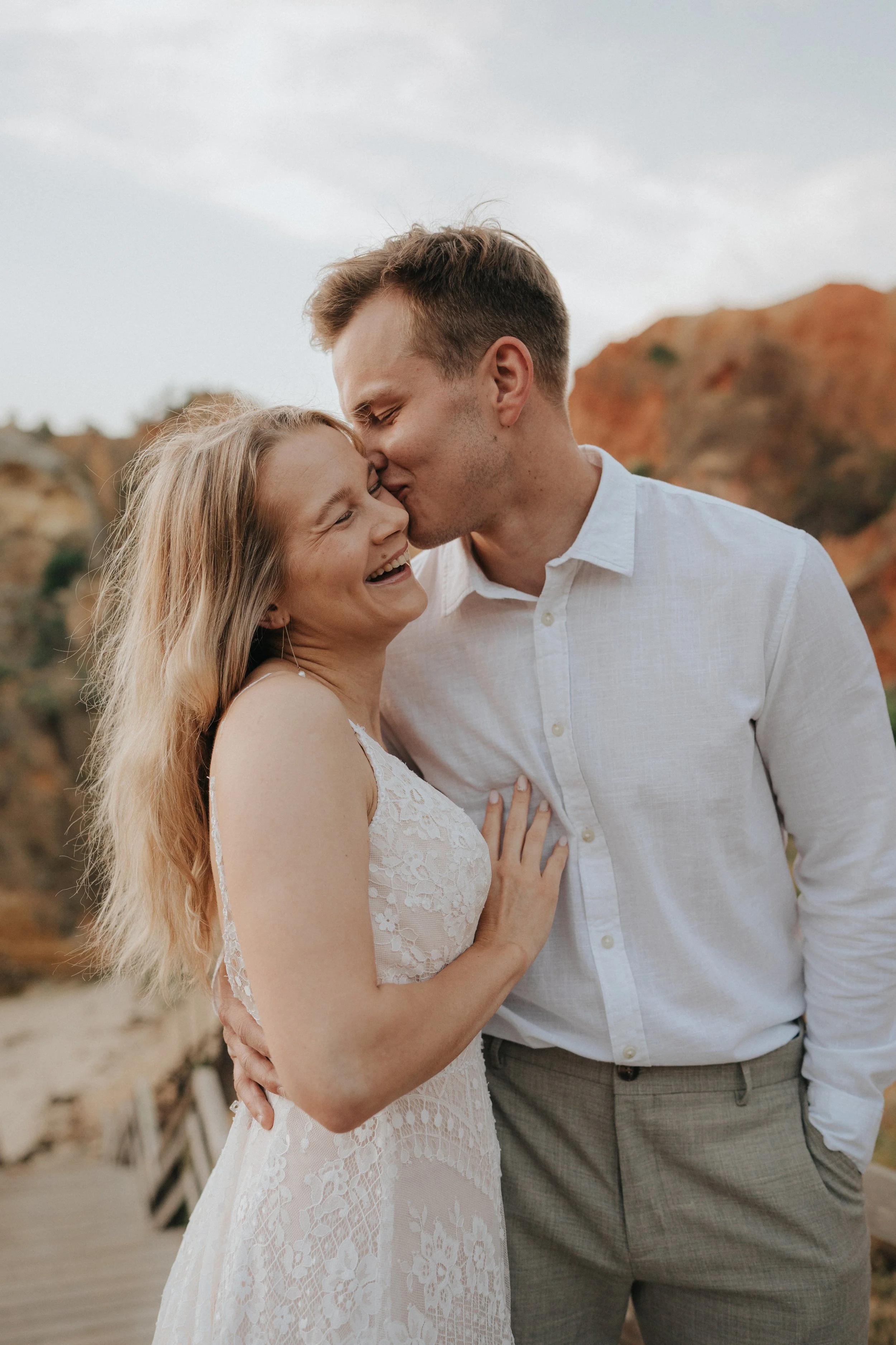A couple sharing a kiss outdoors, with the woman smiling and the man gently kissing her forehead, there are rocky formations in the background.
