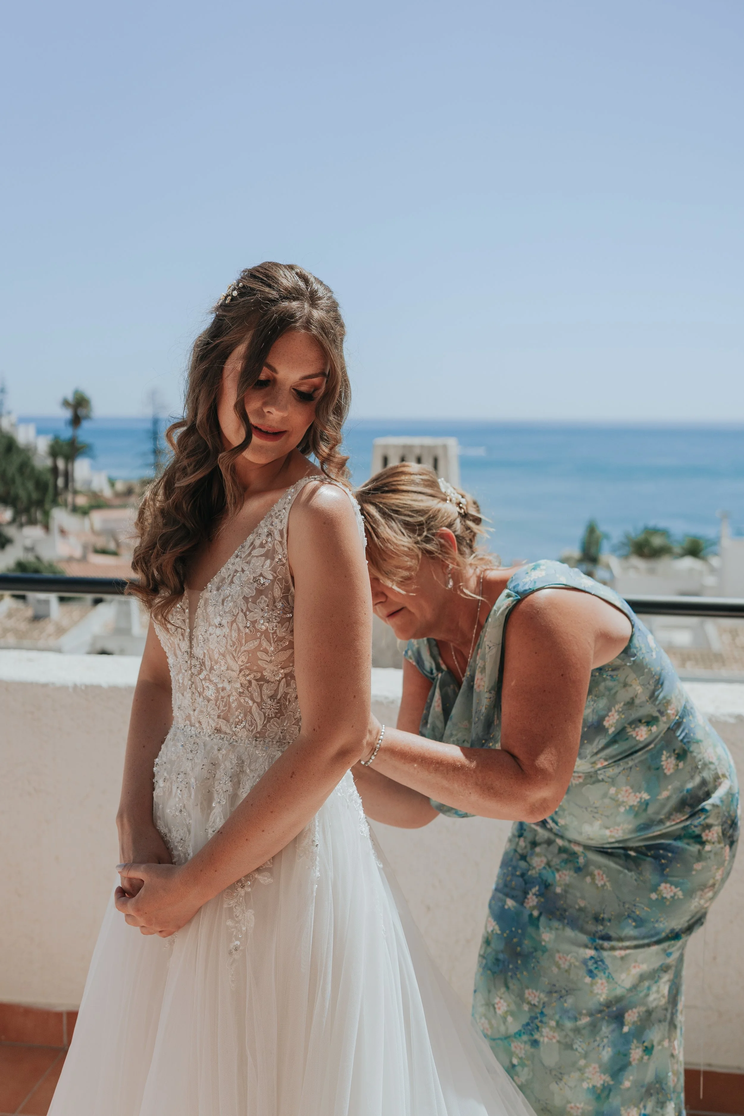 A young woman in a wedding dress is being assisted by an older woman in a floral dress, set against a background of ocean and blue sky.