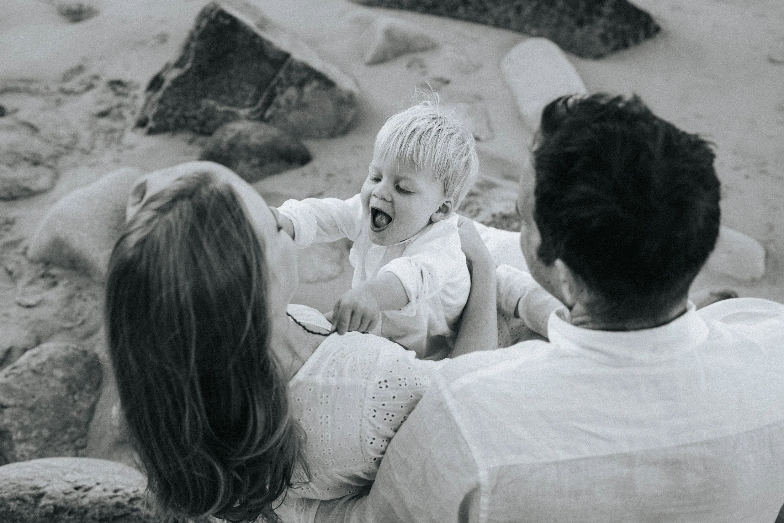 A young blonde boy in a white shirt sitting with adults on a sandy beach, engaging with them and smiling.