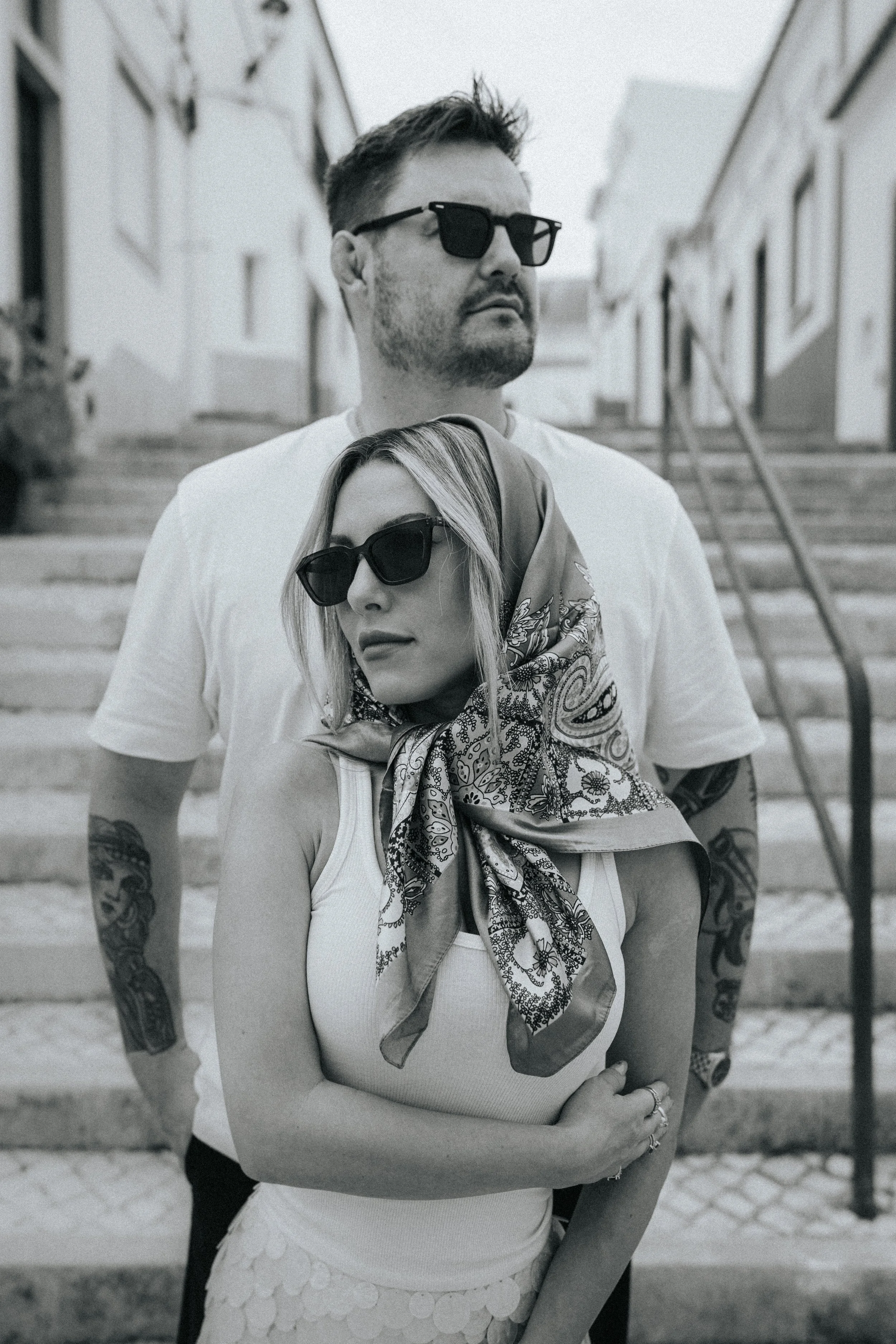 Black and white photo of a man and woman standing outdoors on stairs, both wearing sunglasses. The woman is in front, with a bandana and casual tank top, and the man behind her with short hair and a beard, wearing a t-shirt.