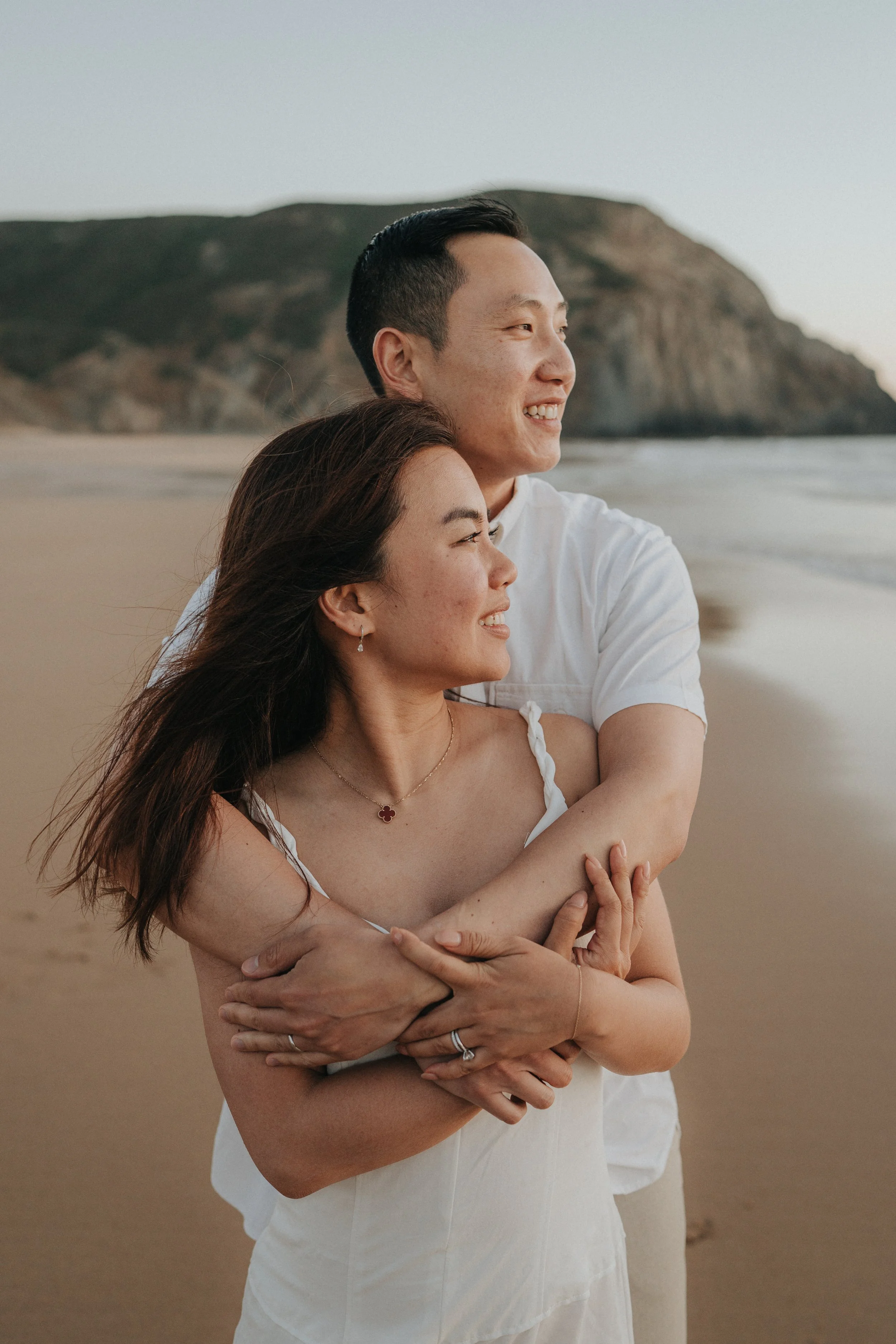 A smiling couple at the beach, with the woman in a white dress and the man in a white shirt, embracing each other with their arms crossed, and a mountain in the background.
