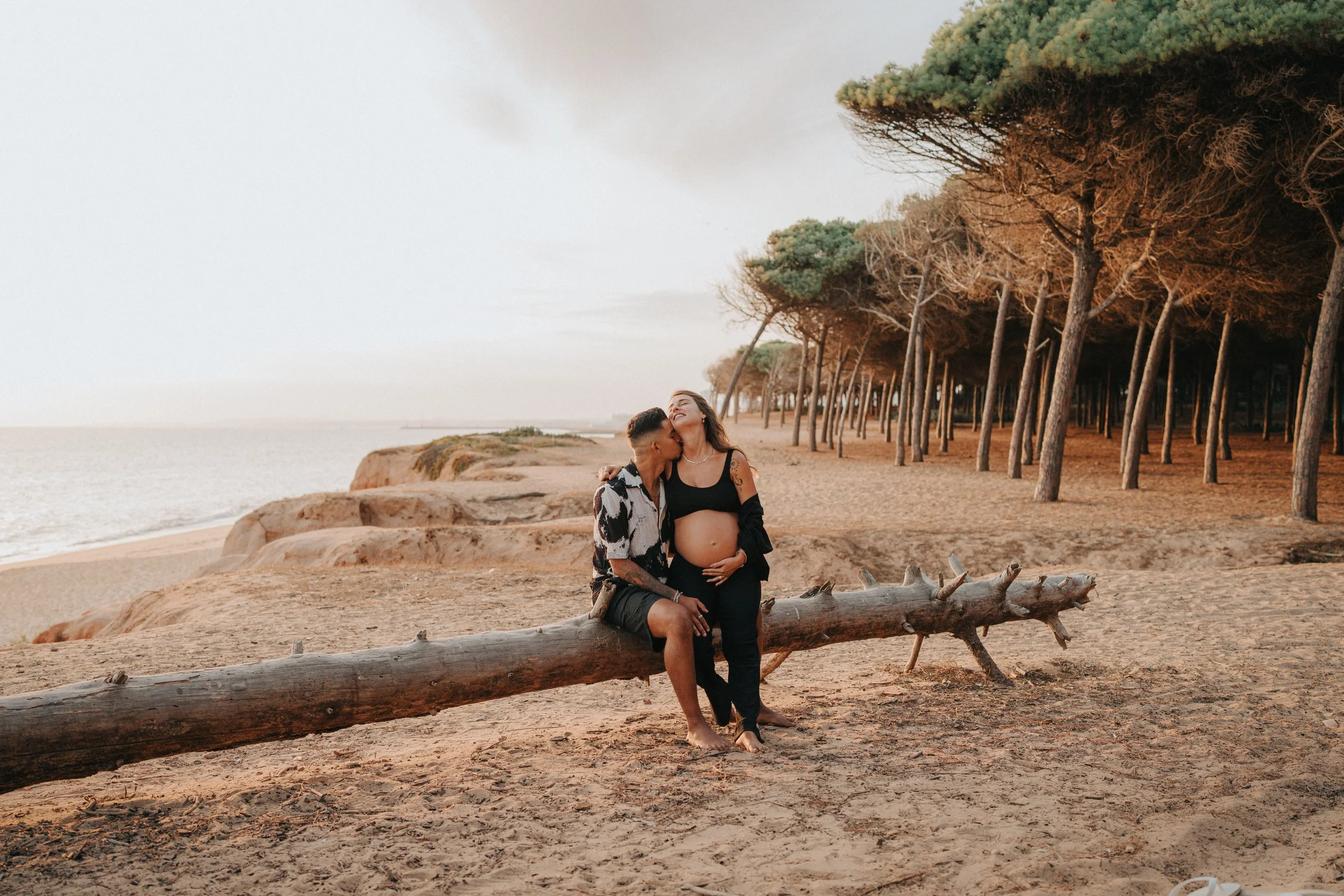 A pregnant woman and a man with tattoos sitting on a log on a sandy beach, kissing and smiling, with a row of trees and the ocean in the background.