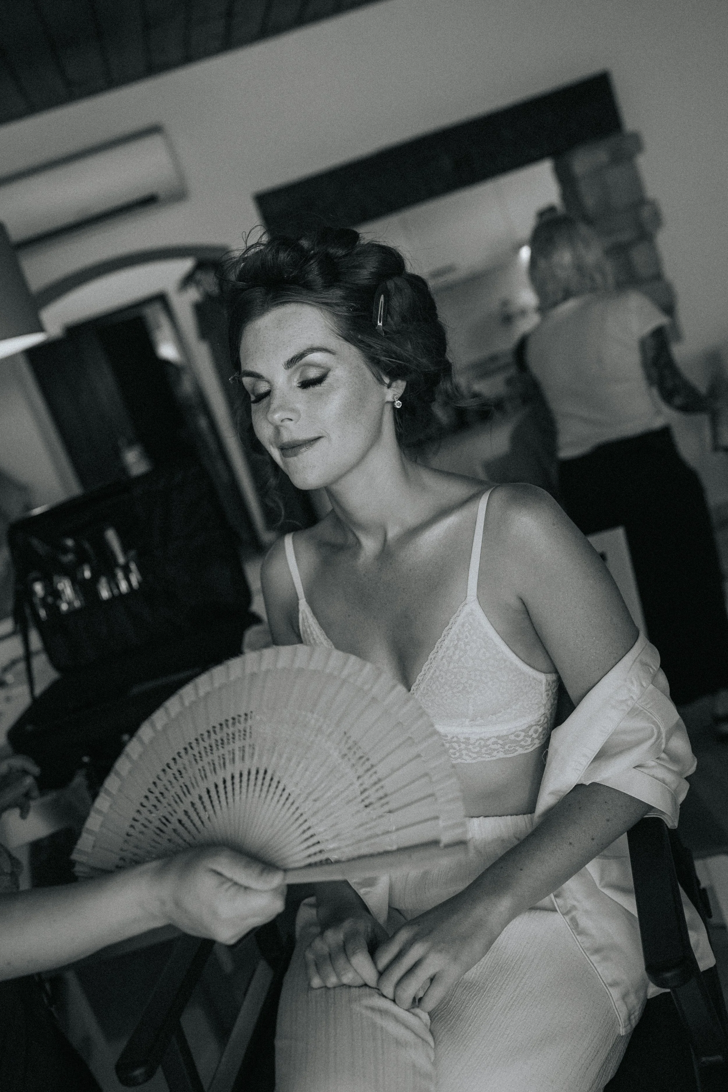 A woman with styled hair and earrings smiling with her eyes closed while holding a fan, in a setting with a woman in the background and other objects.