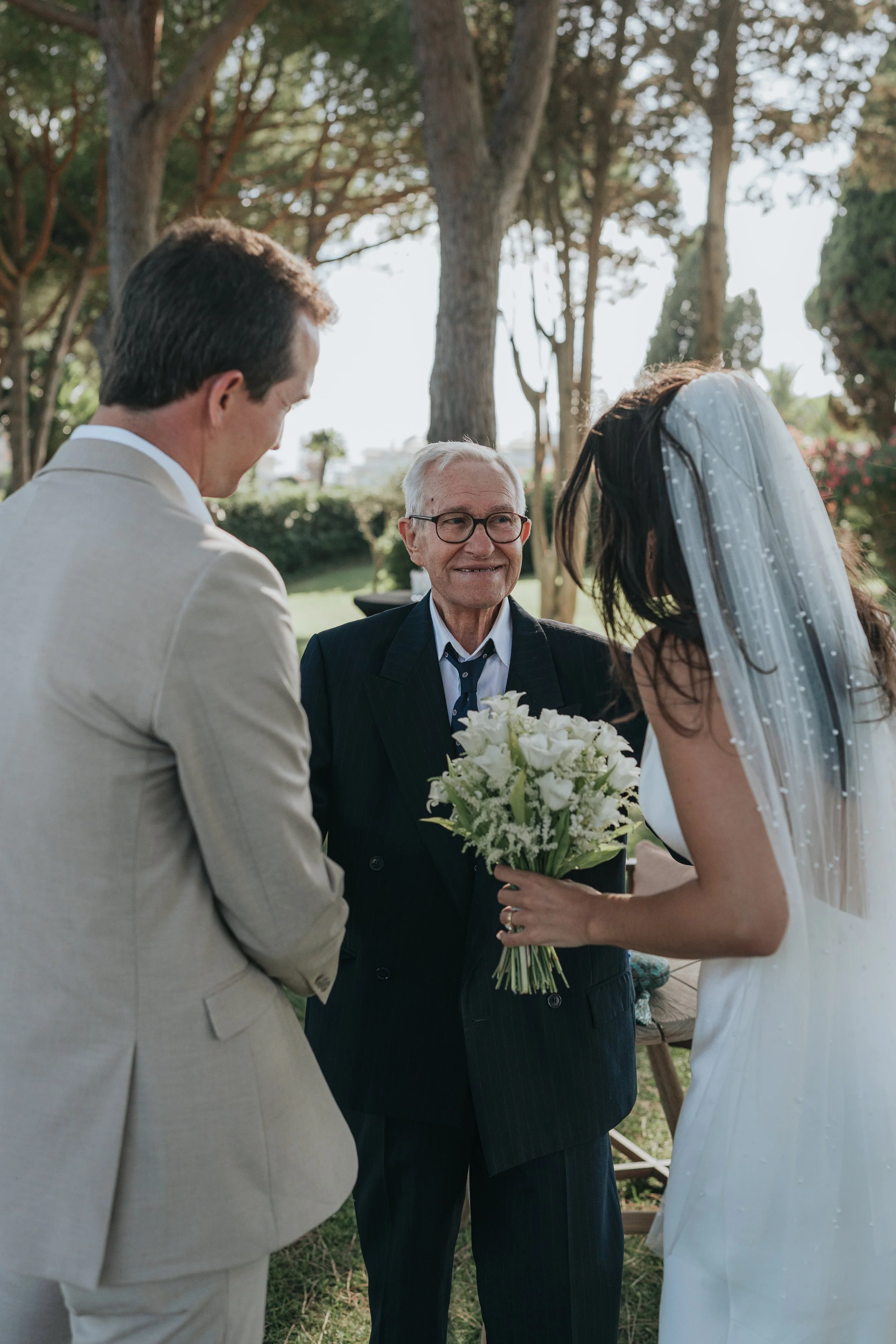 A wedding ceremony outdoors with an officiant, a bride, and a groom. The officiant is smiling while holding a bouquet of white flowers. The bride, in a white dress and veil, is leaning to speak. The groom, in a light-colored suit, is facing the coupl