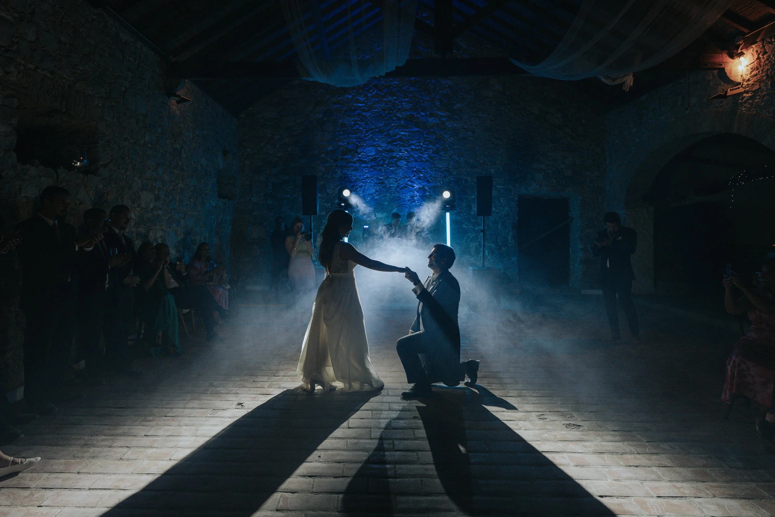 A couple getting married in a rustic, stone-walled venue with dim lighting and a foggy atmosphere. The bride is reaching out to the groom, who is kneeling, during a dance or ceremony, with wedding guests watching.