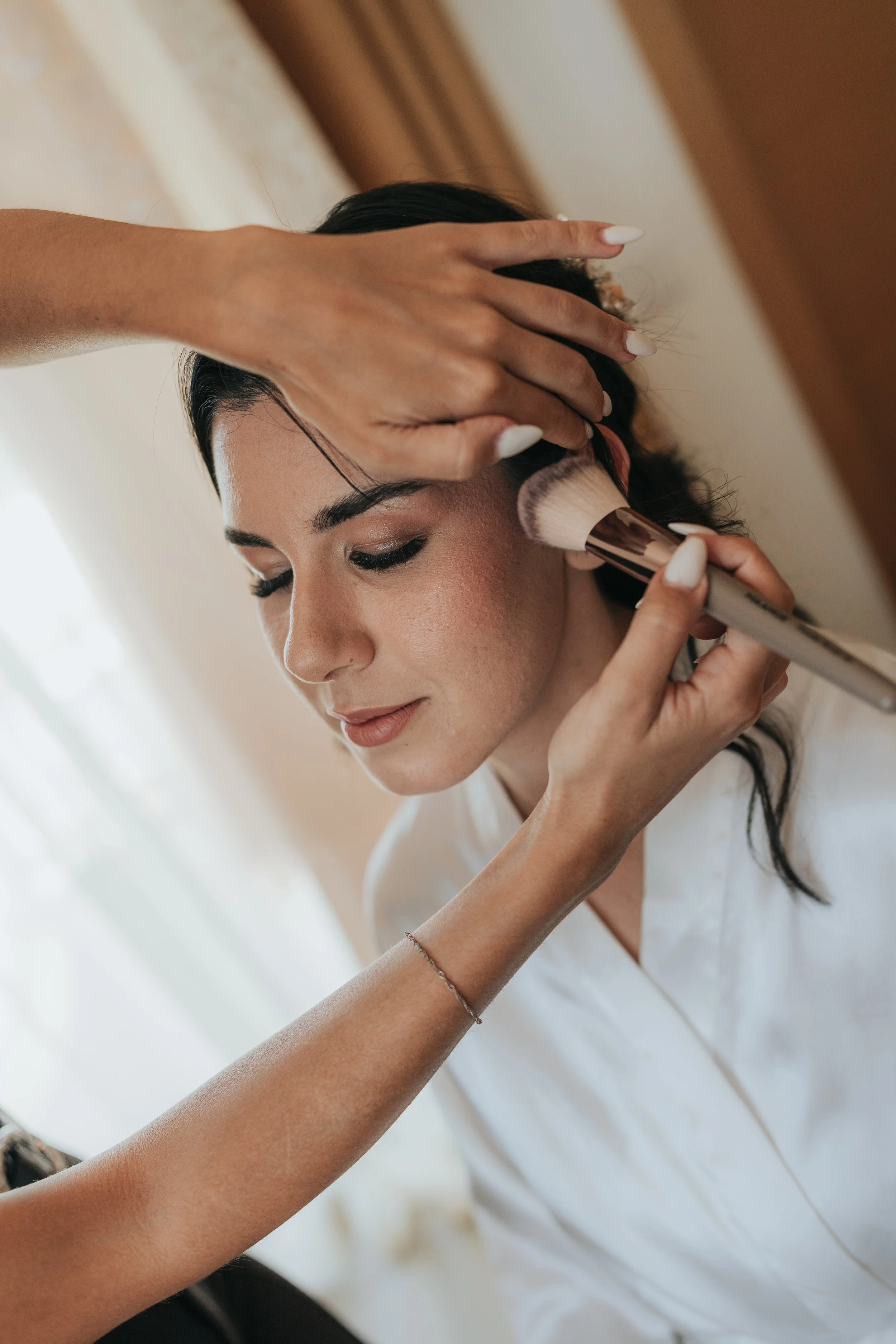 A woman with dark hair is having makeup applied by another person with a brush, with her eyes closed and head slightly tilted. The person applying makeup has long nails painted white.