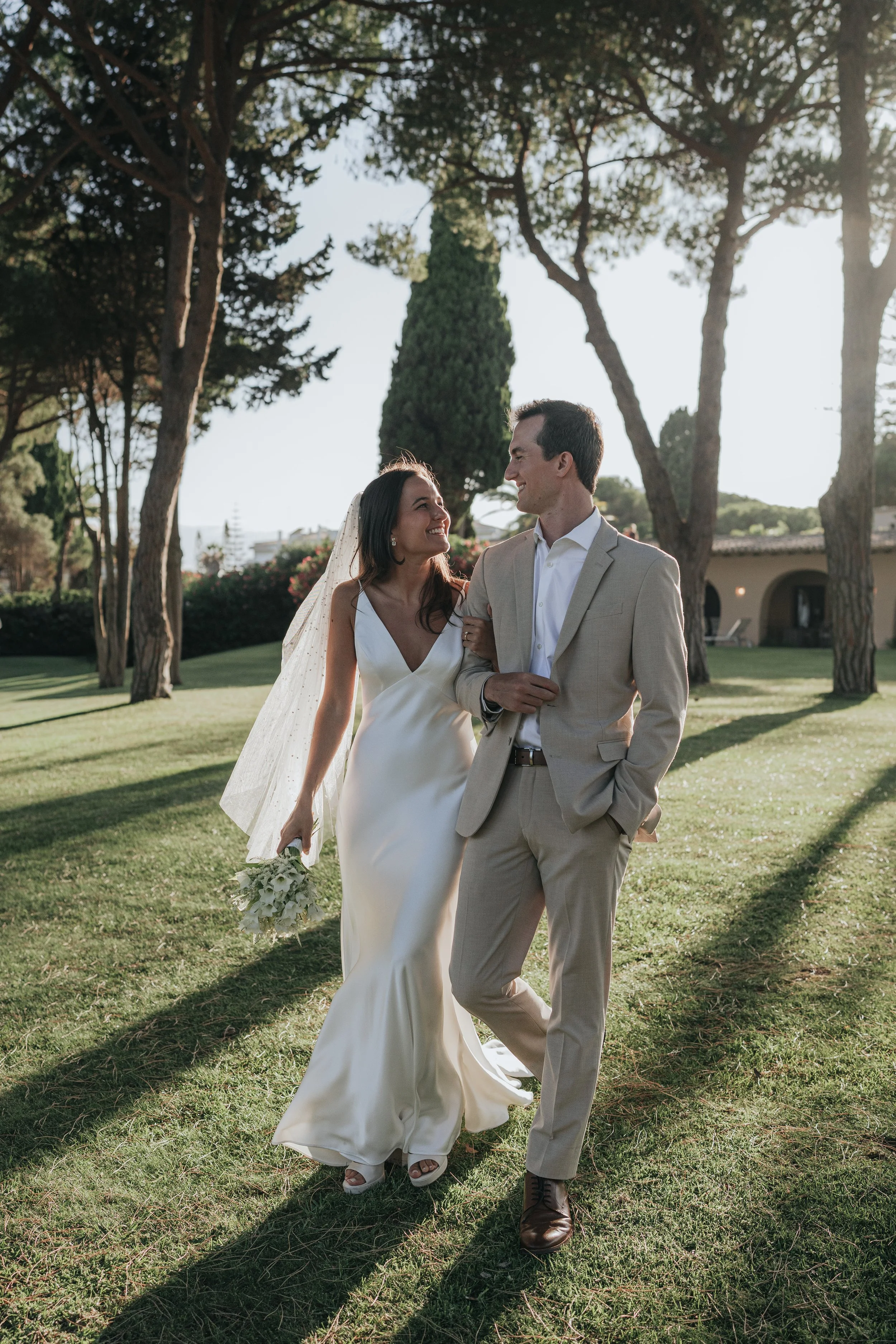 A bride and groom walking arm in arm outdoors in a park-like setting with tall trees, green grass, and a building in the background, enjoying a sunny day after their wedding.
