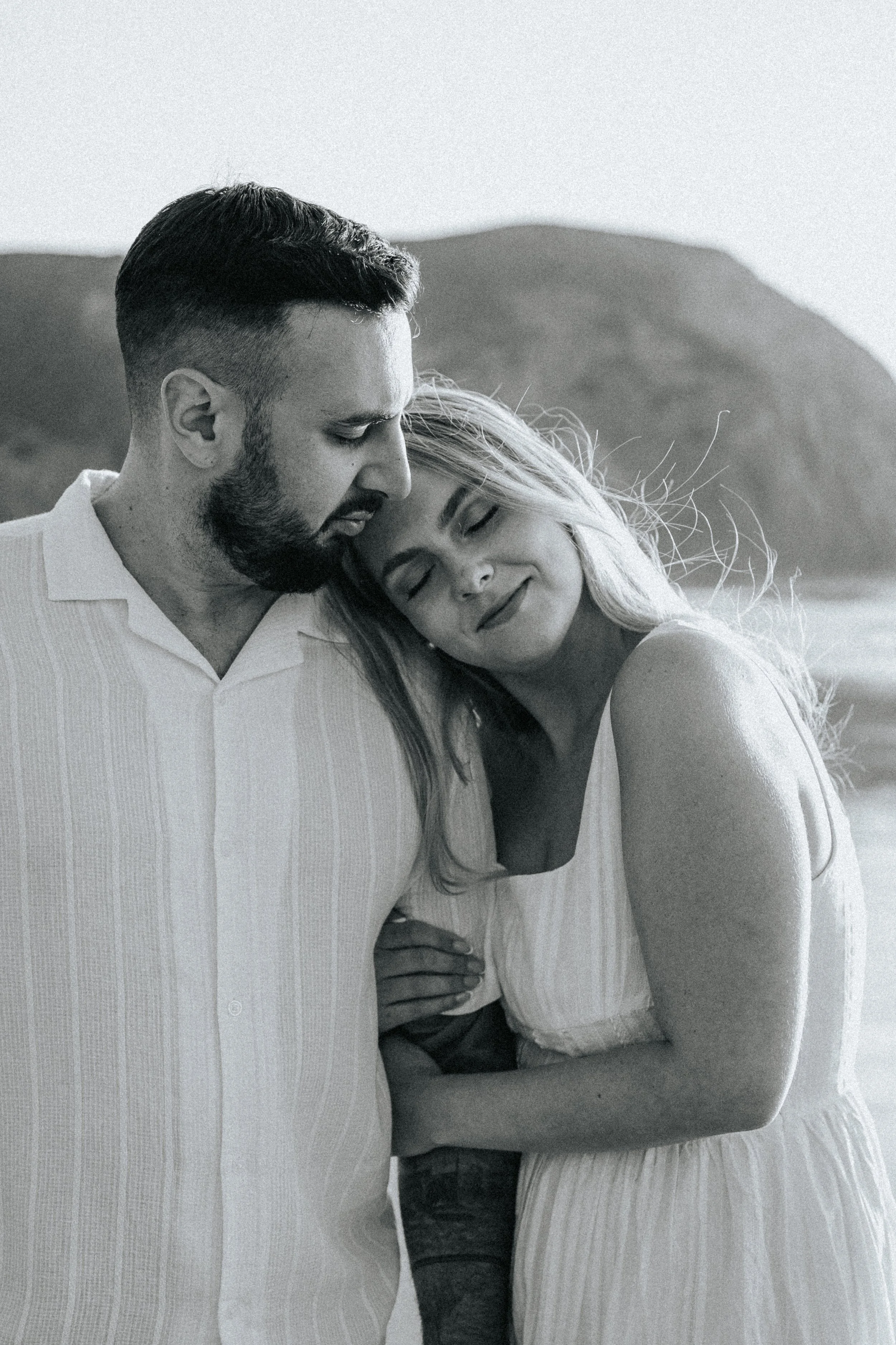 A man and a woman are embracing each other outdoors with mountains in the background. The woman rests her head on the man's shoulder and both are smiling softly. The image is in black and white.