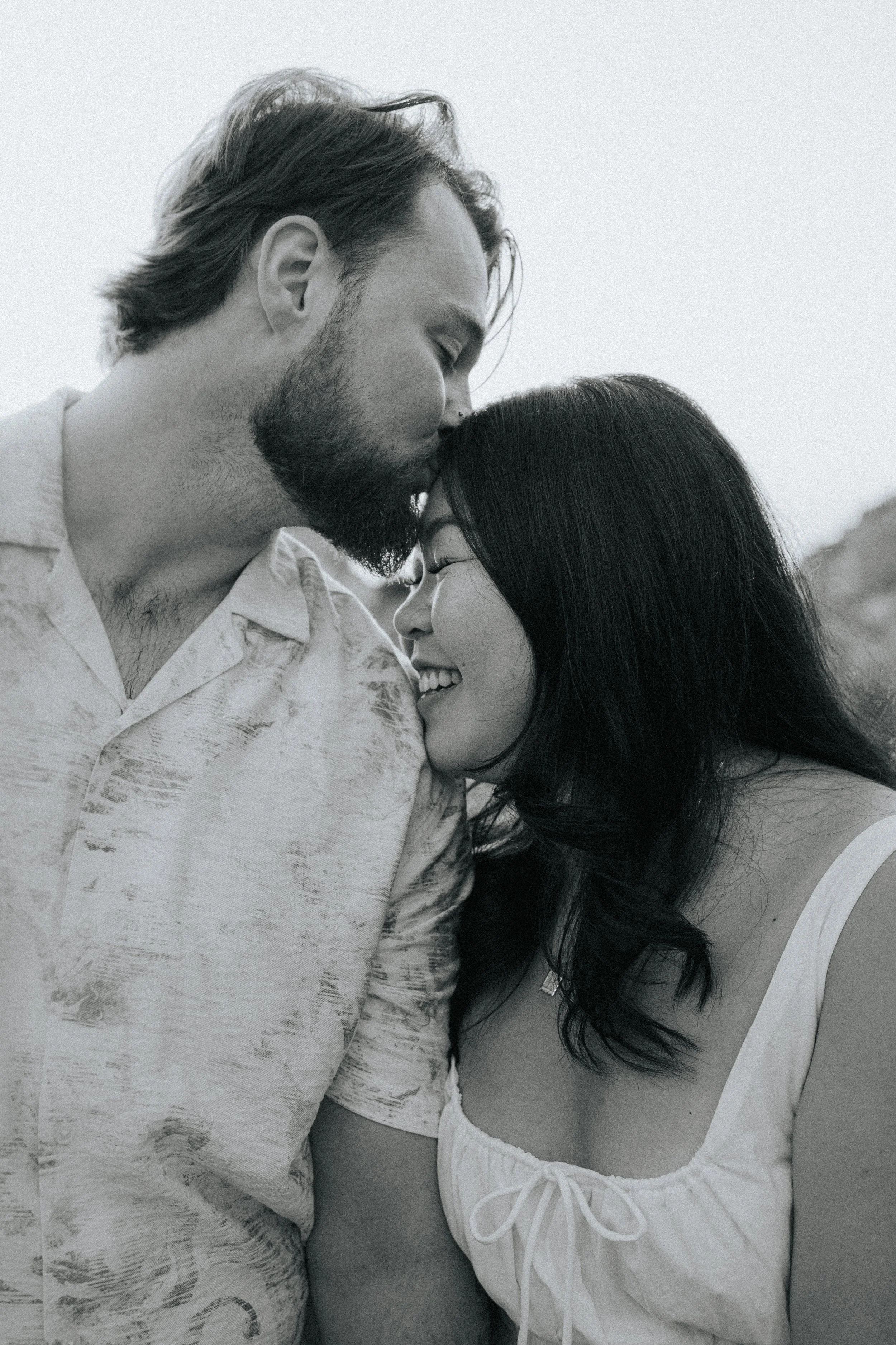 A black and white photo of a couple touching foreheads and smiling, outdoors with natural light.