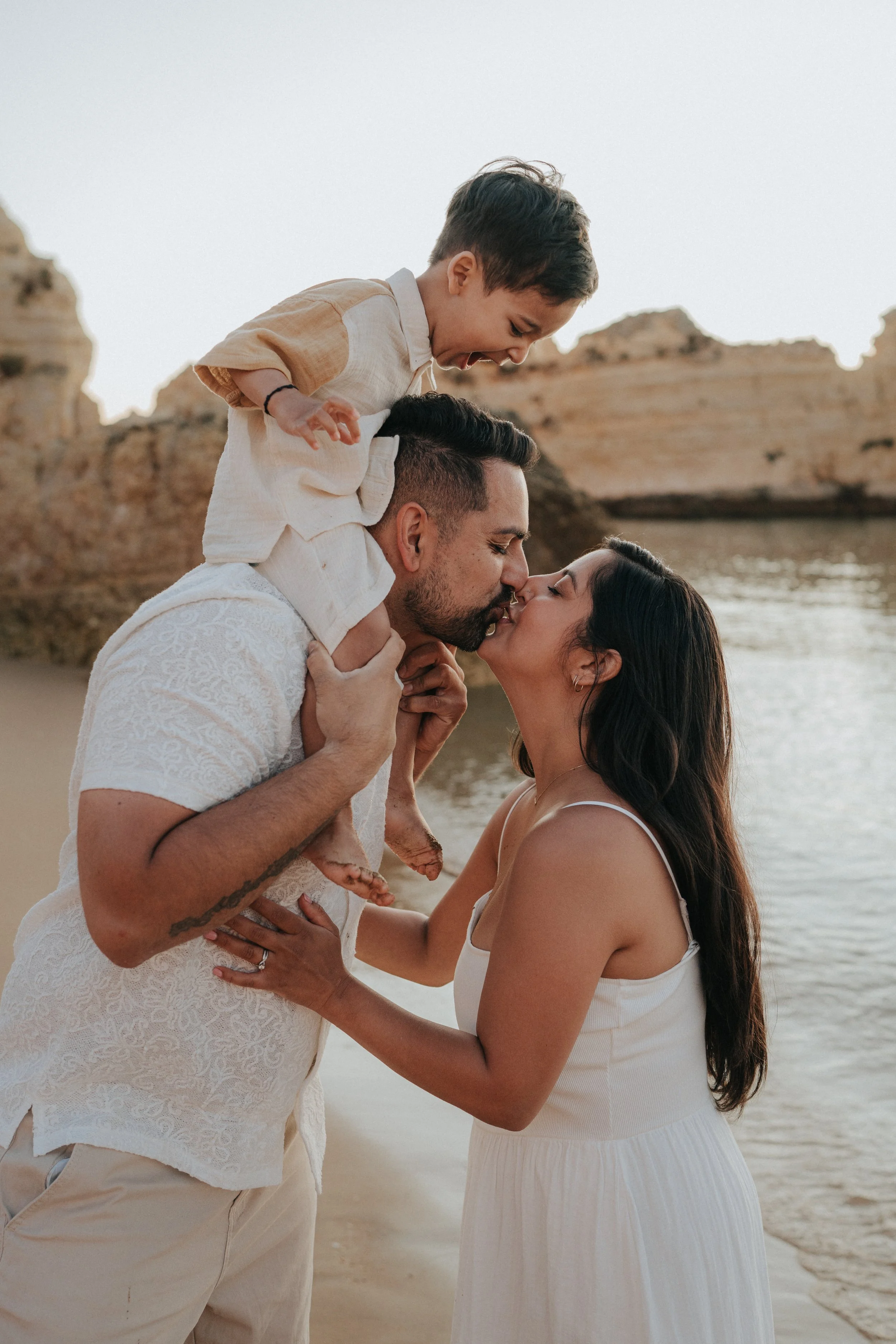 A couple and a young boy on the beach, the couple kissing and the boy smiling, with rocks and water in the background during sunset.