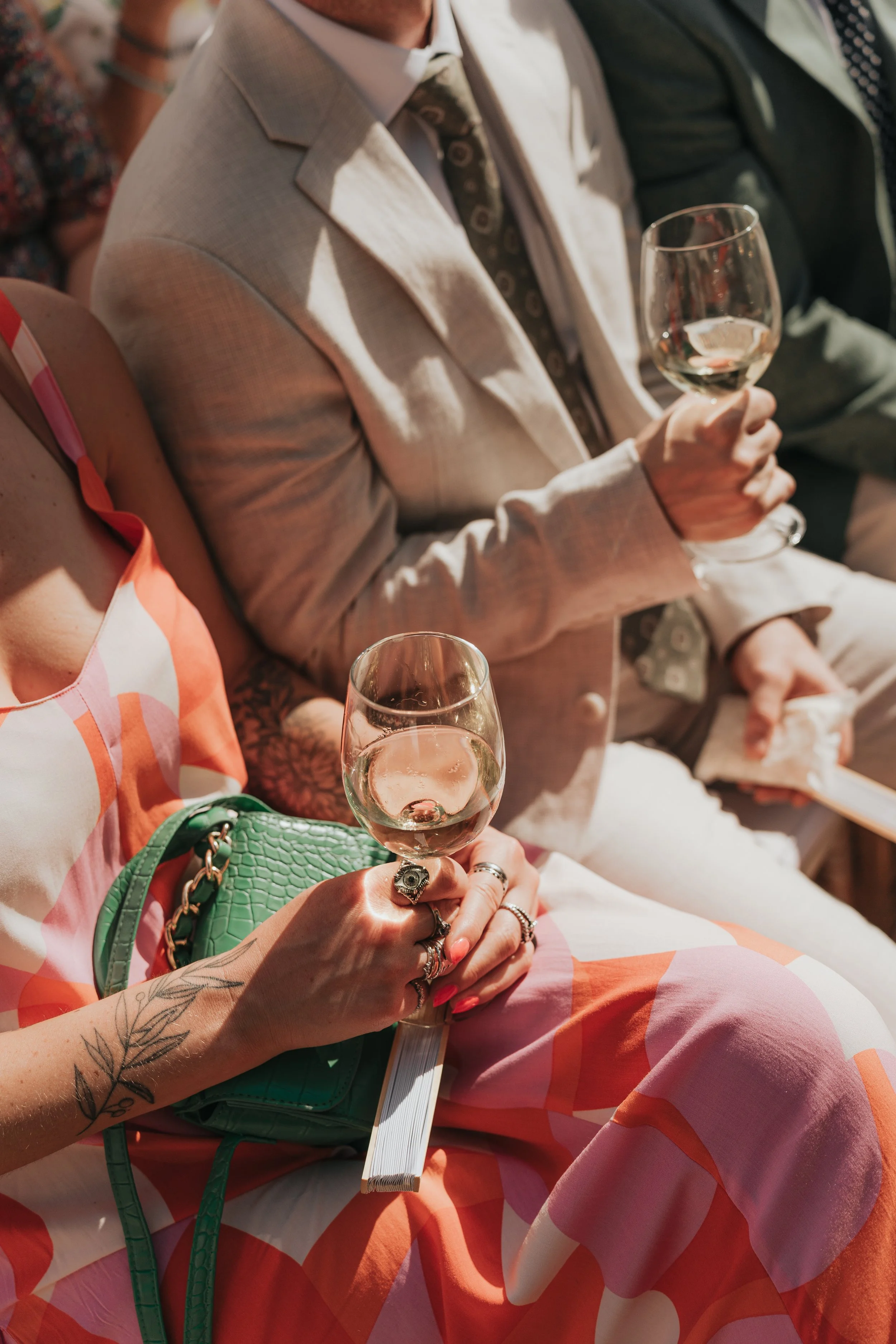 A woman holding a glass of white wine during a social event, wearing a colorful dress, with a tattoo on her arm and a green purse on her lap. Other people are visible in the background.