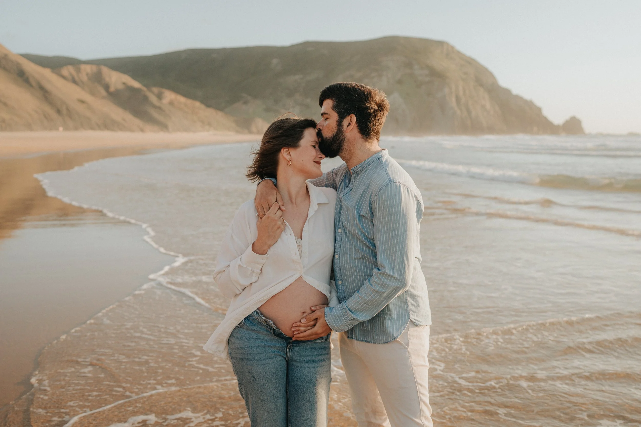 A man and woman are standing on a beach, embracing each other with the man kissing the woman's forehead. The woman is pregnant and smiling with her eyes closed. They are near the shoreline with waves and cliffs in the background.