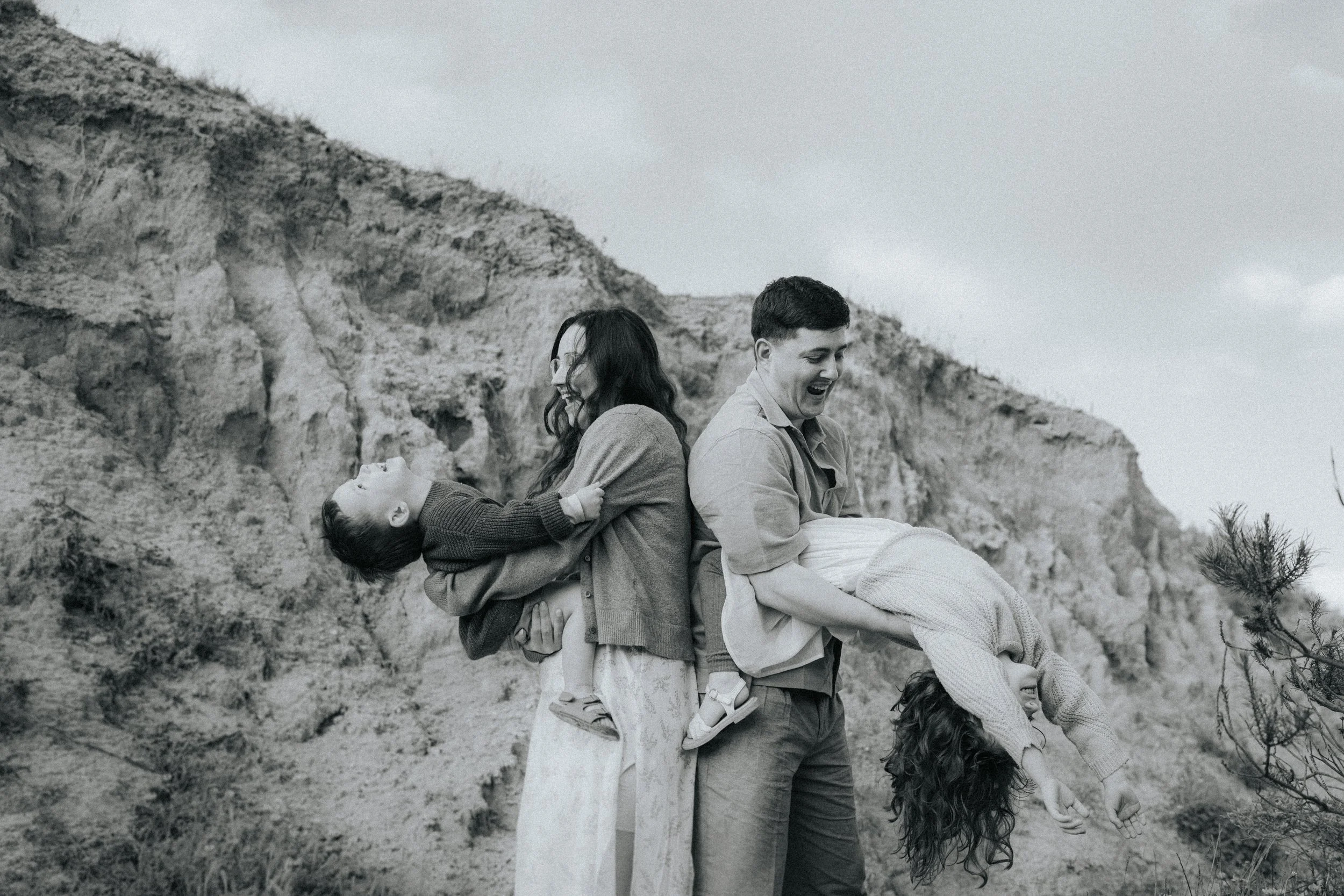 A black and white photo of a group of young people with three girls and one boy, playing and smiling outdoors near a rocky hillside with trees and cloudy sky.