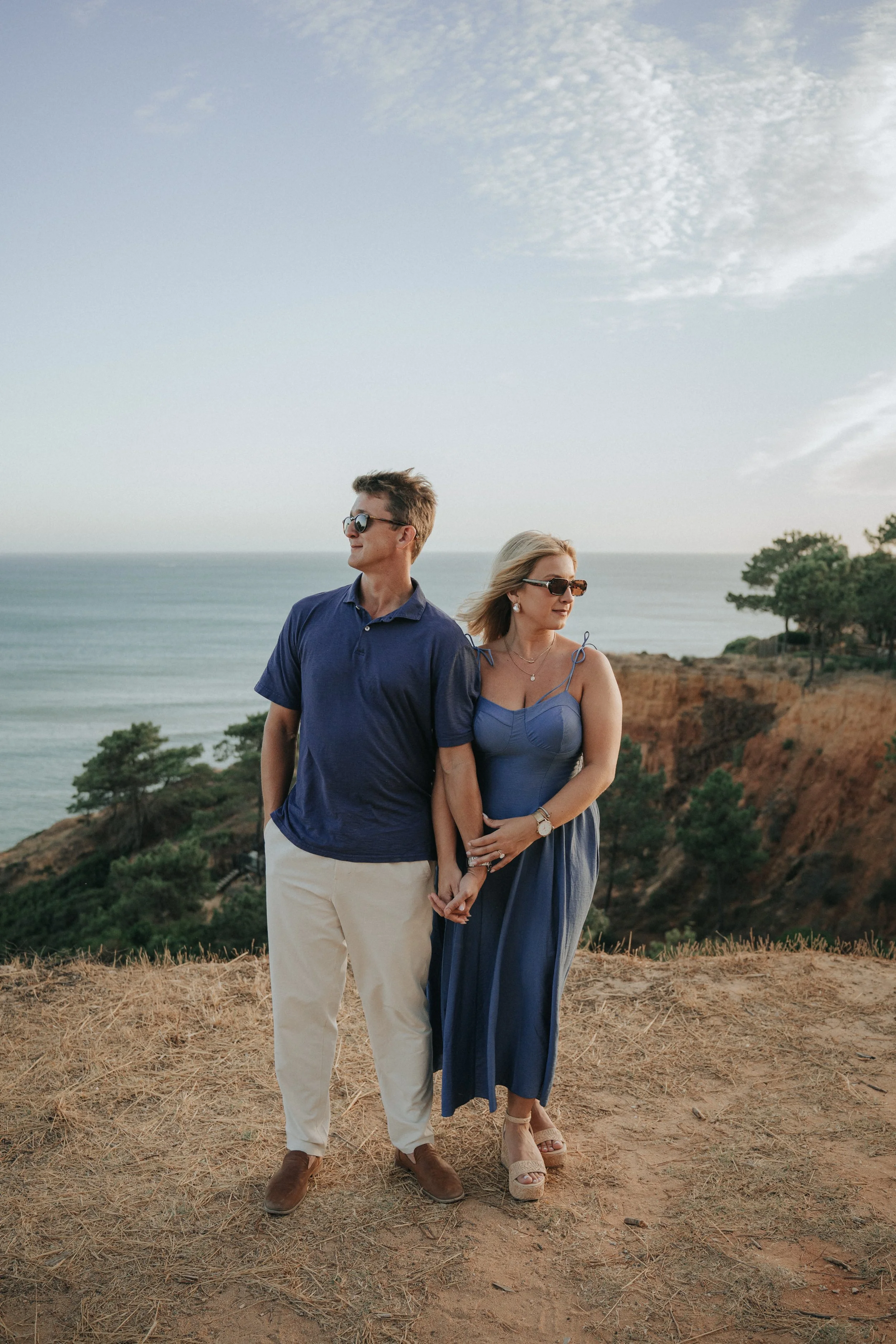 A man and woman standing close together outdoors near the coast, holding hands, with the ocean, sky, and cliffs in the background during daylight.