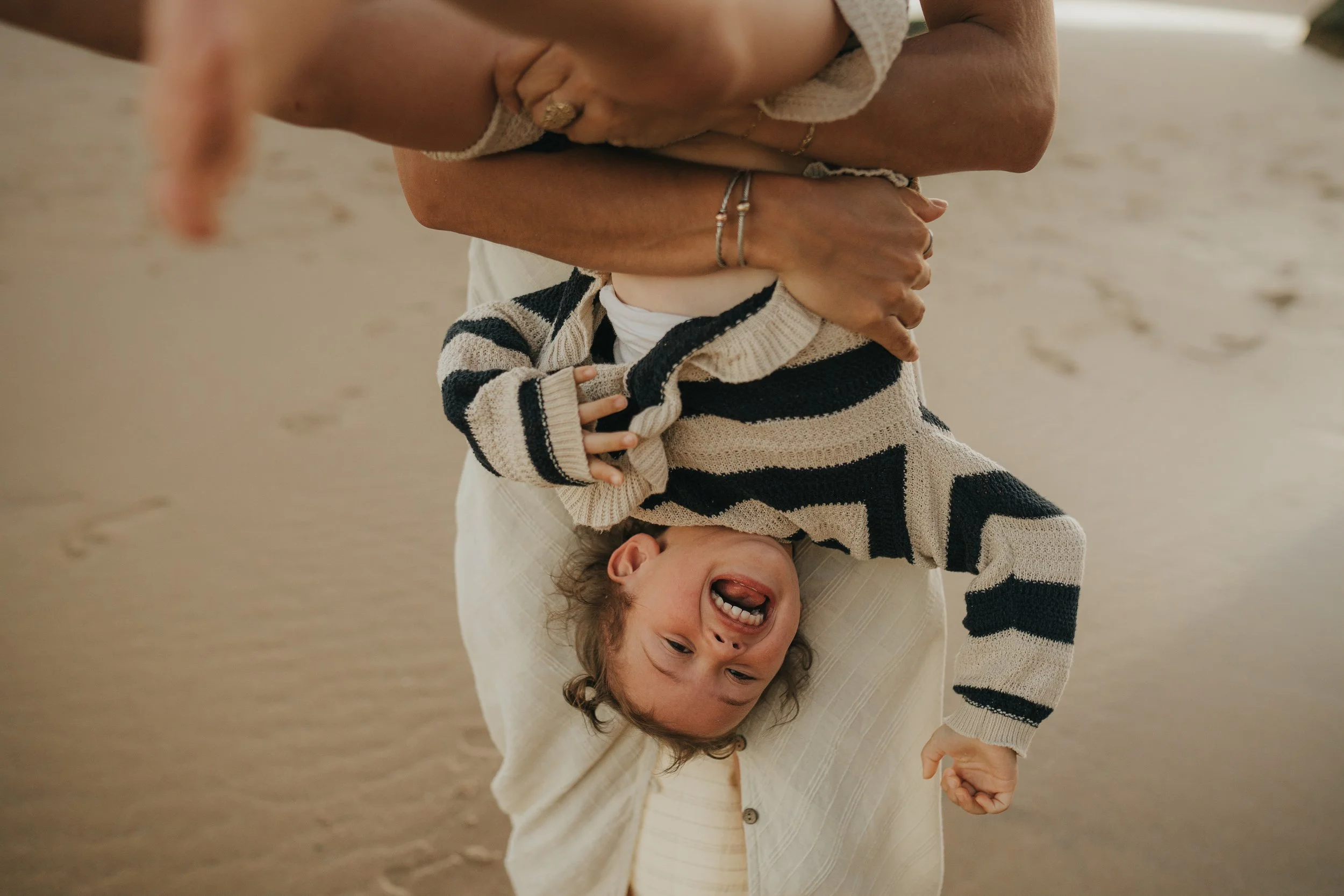 A woman holding a laughing child upside down in a beach setting.