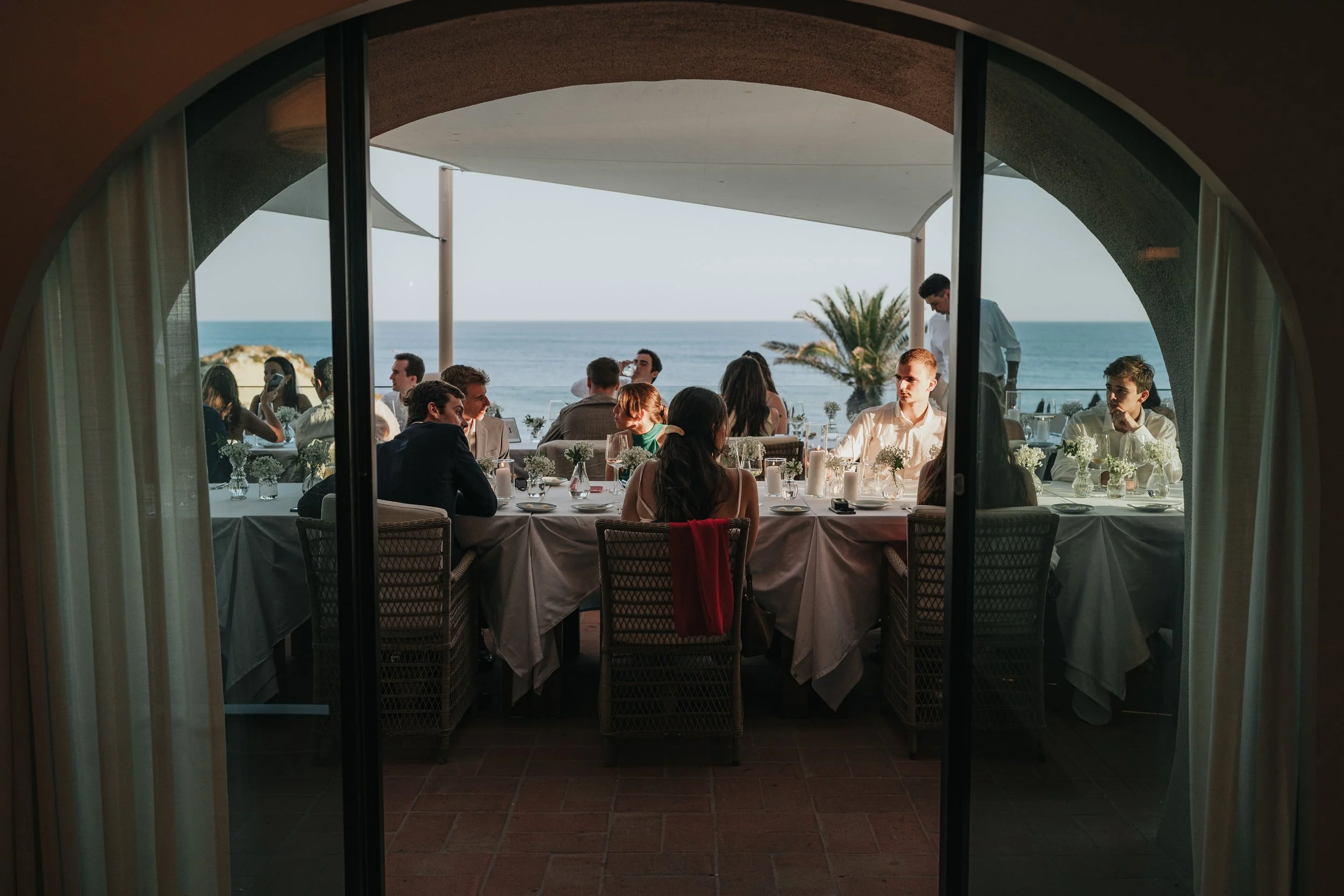 View of a restaurant with large windows overlooking the ocean. Inside, a long table is set with white tablecloths, glassware, candles, and floral centerpieces. Several people are seated at the table, engaged in conversation. The scene is viewed from 