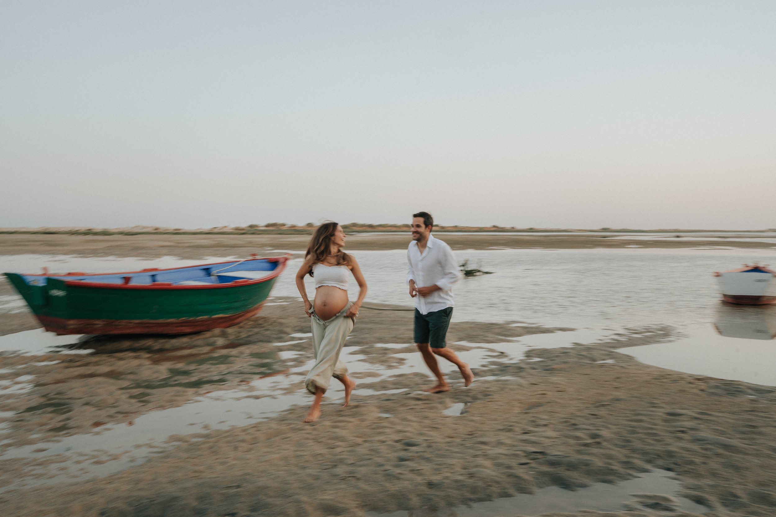 A pregnant woman and a man walk barefoot on a beach near the water, with boats in the background and a cloudy sky.