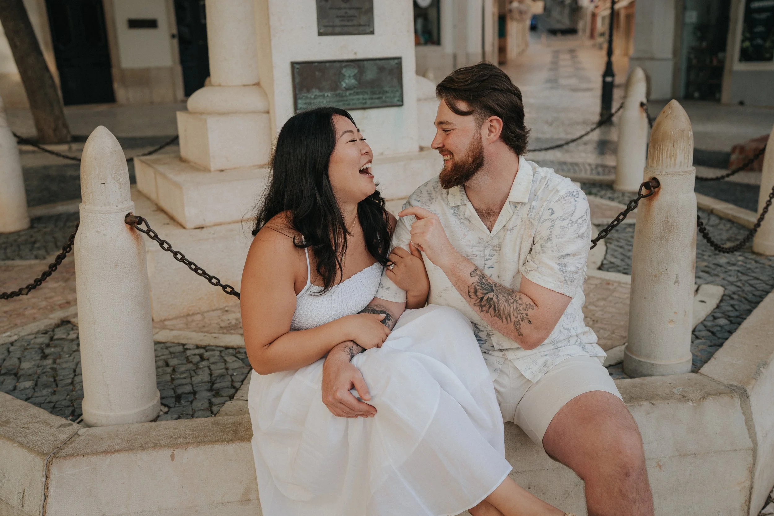 A happy couple sitting on a stone bench in front of a monument, sharing a joyful moment and smiling at each other.