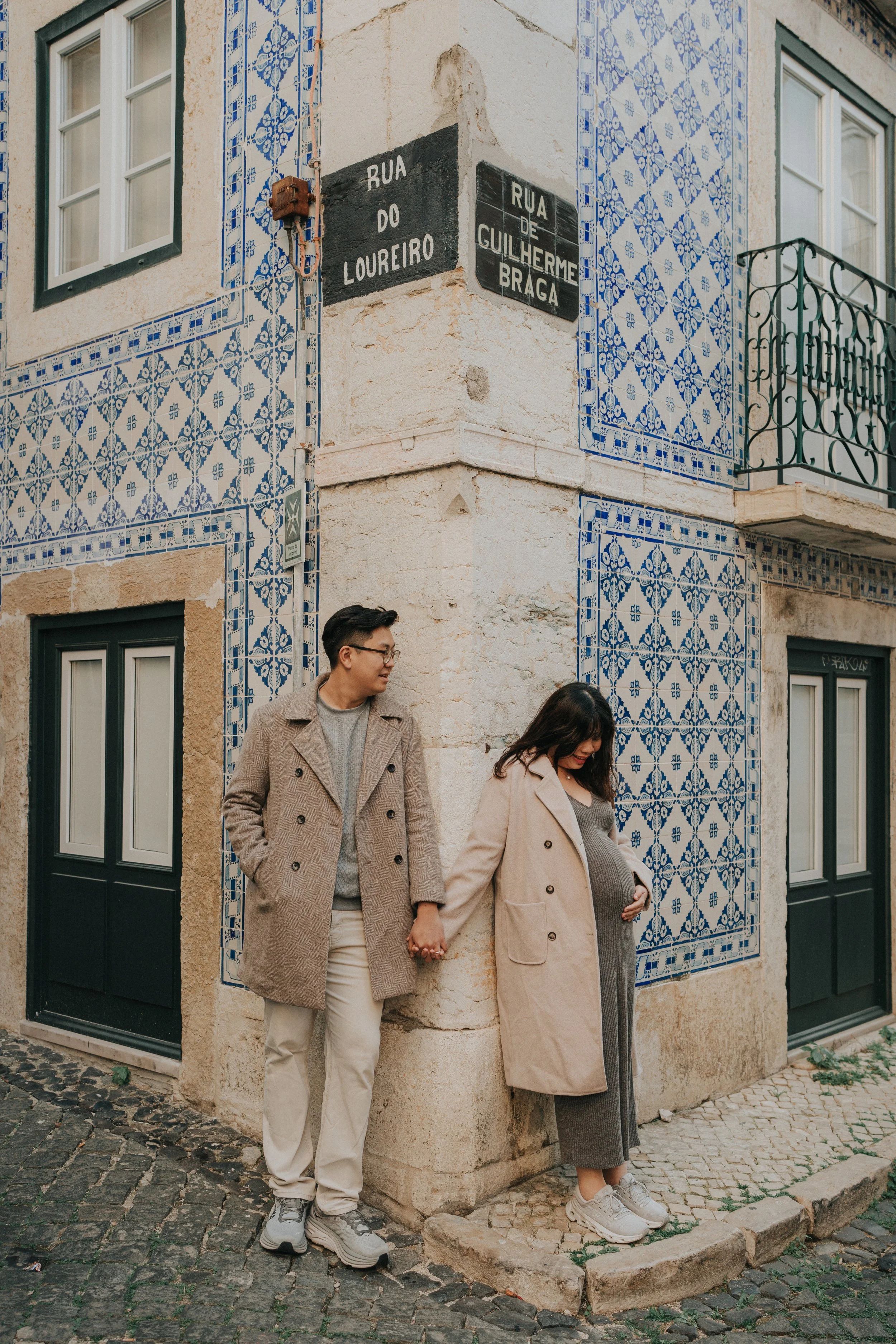 A couple holding hands, standing against the corner of a building with decorative blue and white tiles, in an urban setting with cobblestone street.