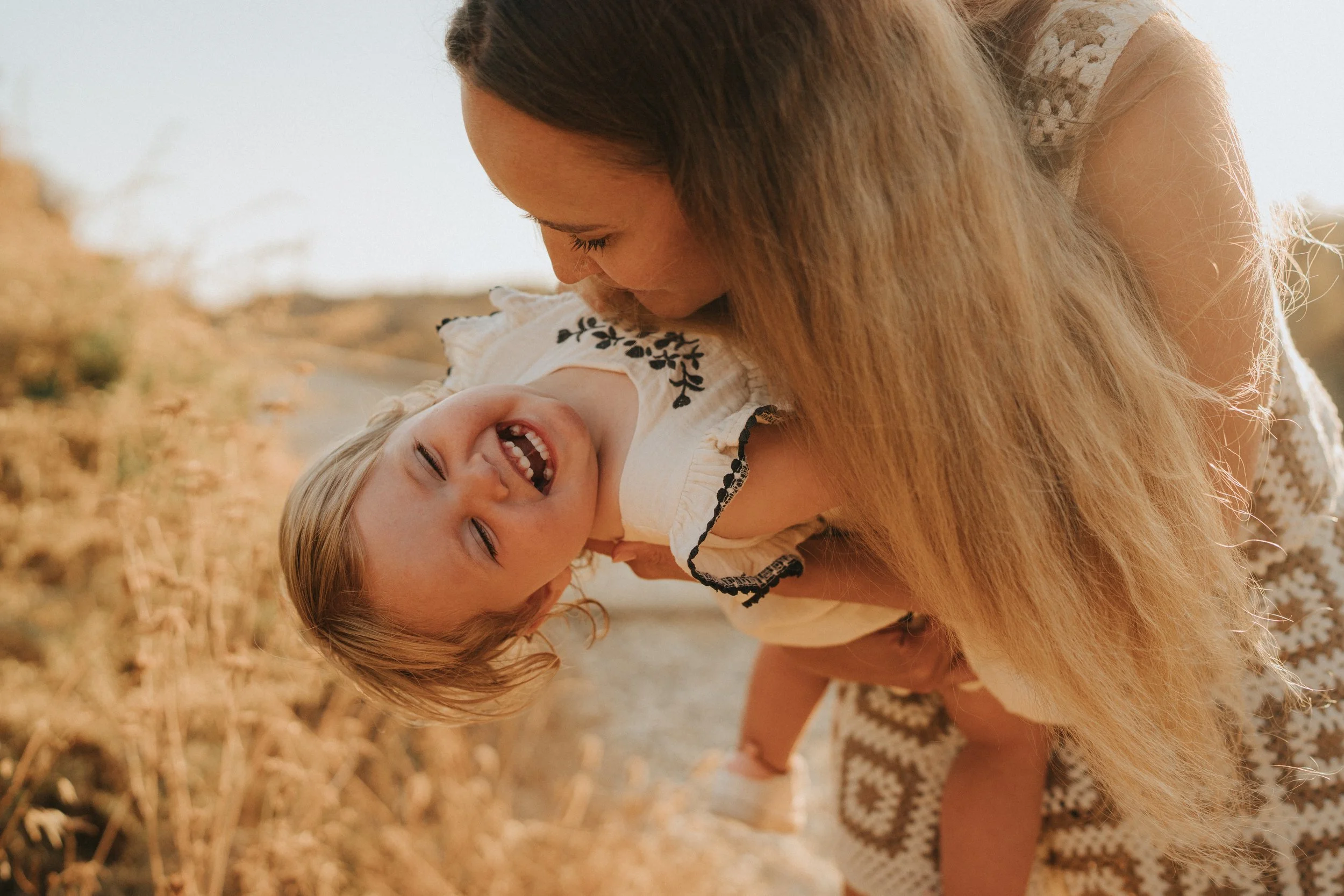 A woman and a young girl outdoors, smiling and laughing, with the woman leaning over the girl as they enjoy a moment together in a natural setting.