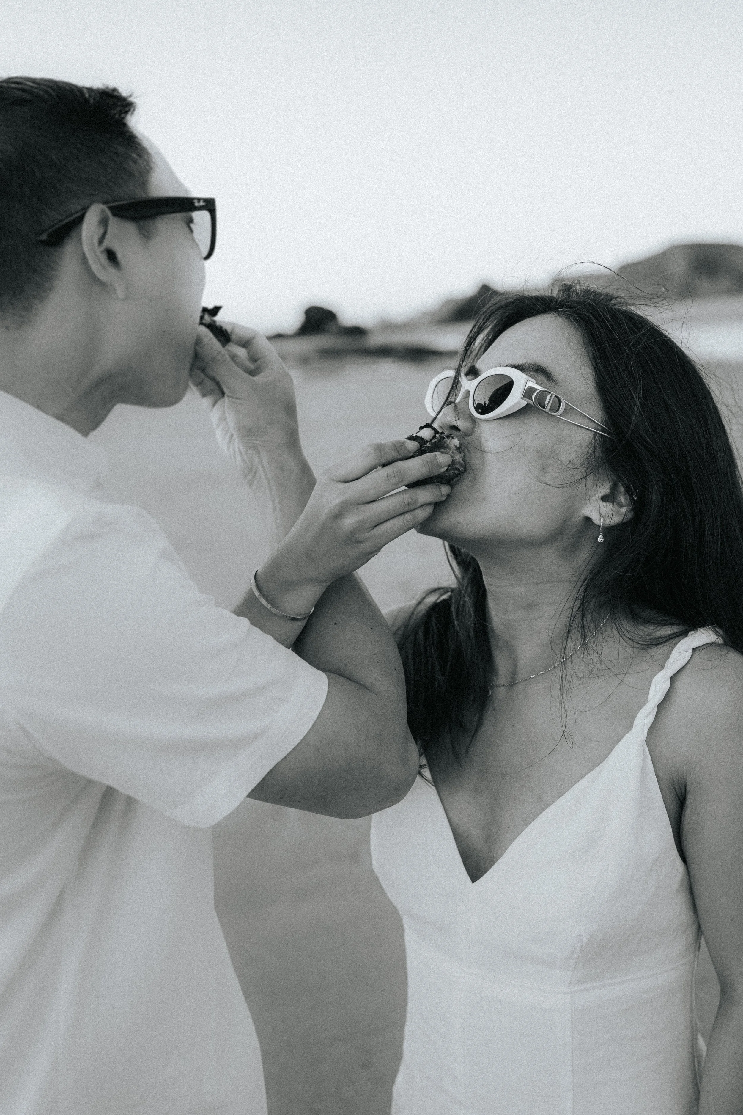 A person is feeding another person a piece of food on a beach, both wearing sunglasses and light-colored clothing.