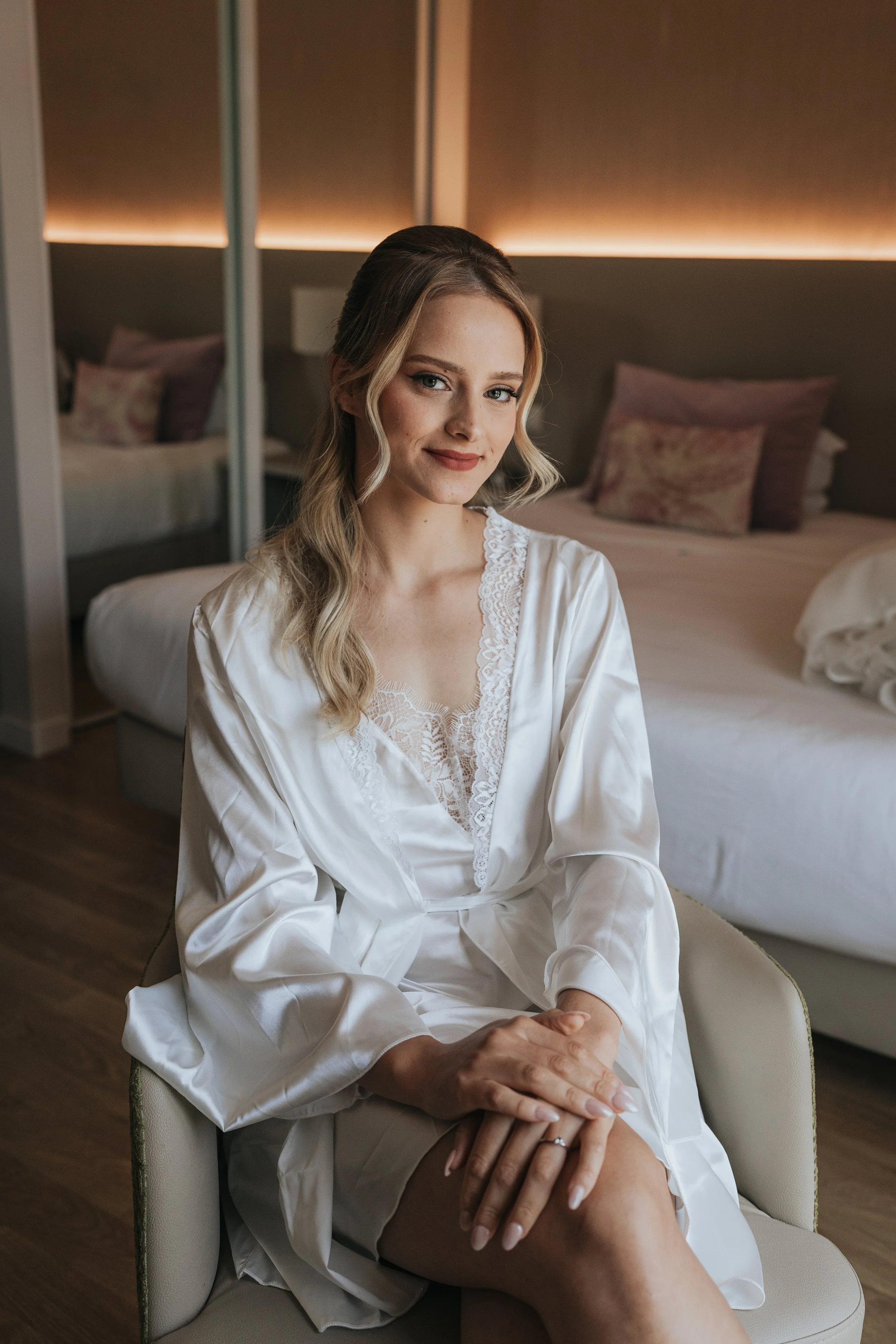 A woman with blonde hair styled in loose waves, wearing a white satin robe, sitting on a chair in a bedroom with beds and floral pillows in the background.