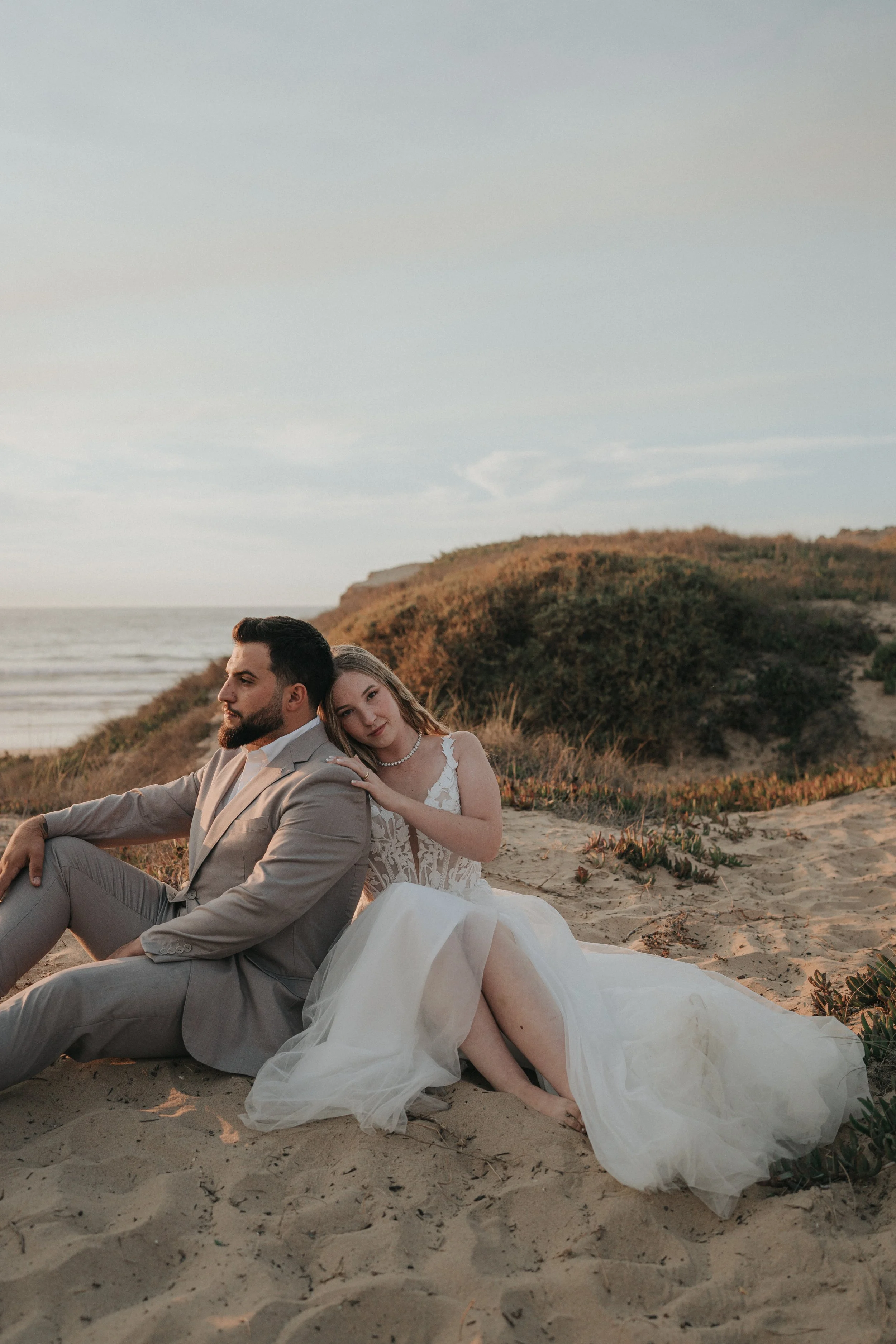 A man in a gray suit sitting on the sand with a woman in a white wedding dress resting her head on his shoulder at the beach during sunset.