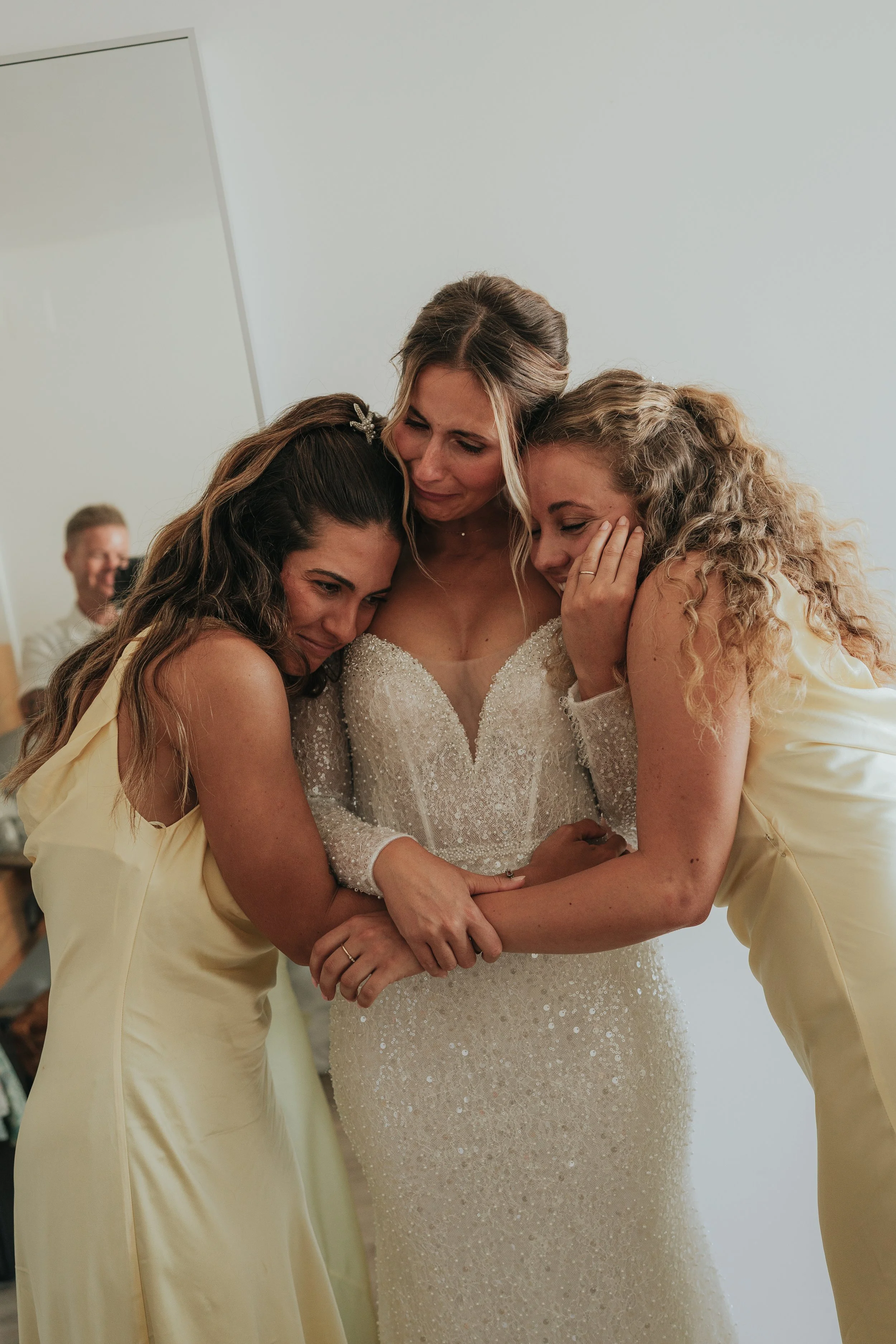 Three women hugging, with the bride in a white wedding dress and two bridesmaids in light yellow dresses, sharing an emotional moment.