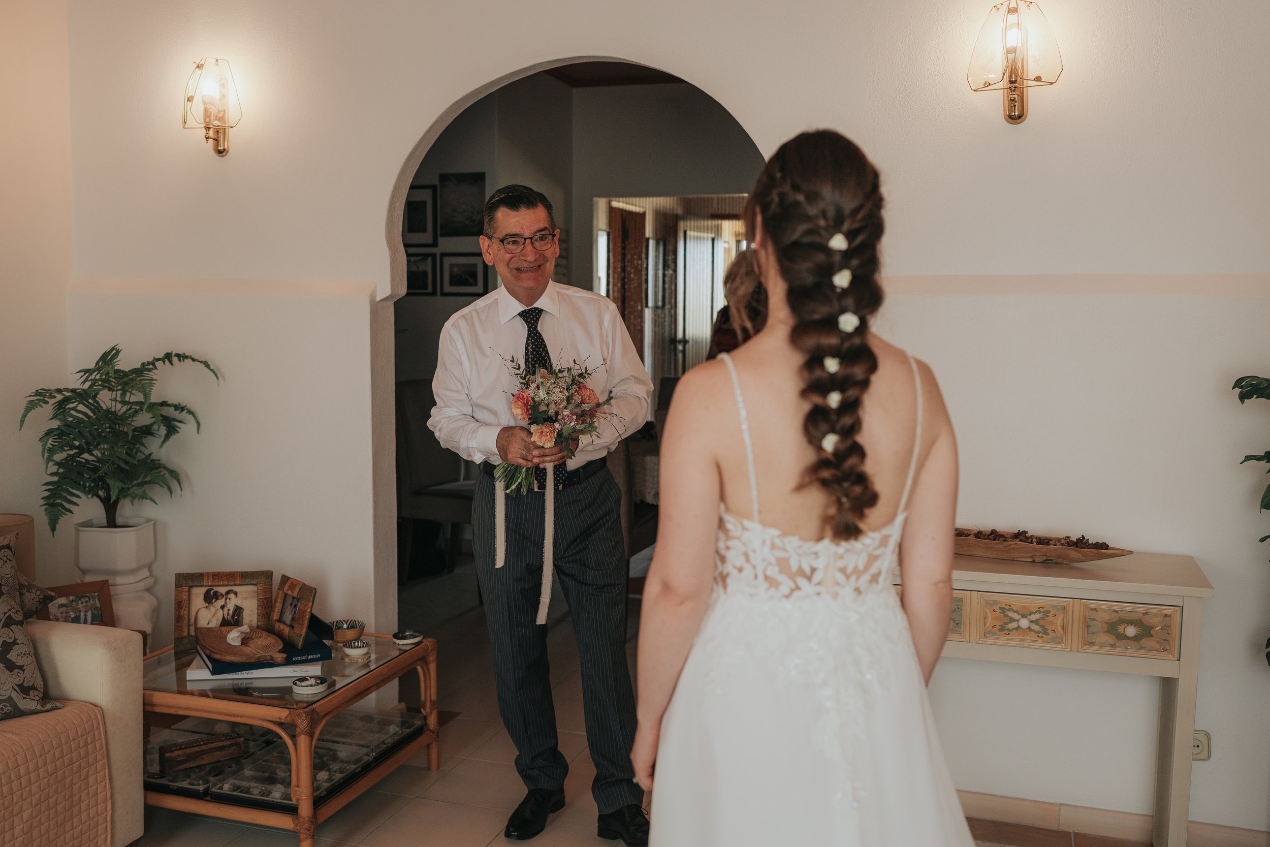 A man in a white shirt and tie holding a bouquet of flowers at the doorway of a home, smiling at a woman in a white lace dress with floral hair accessories.