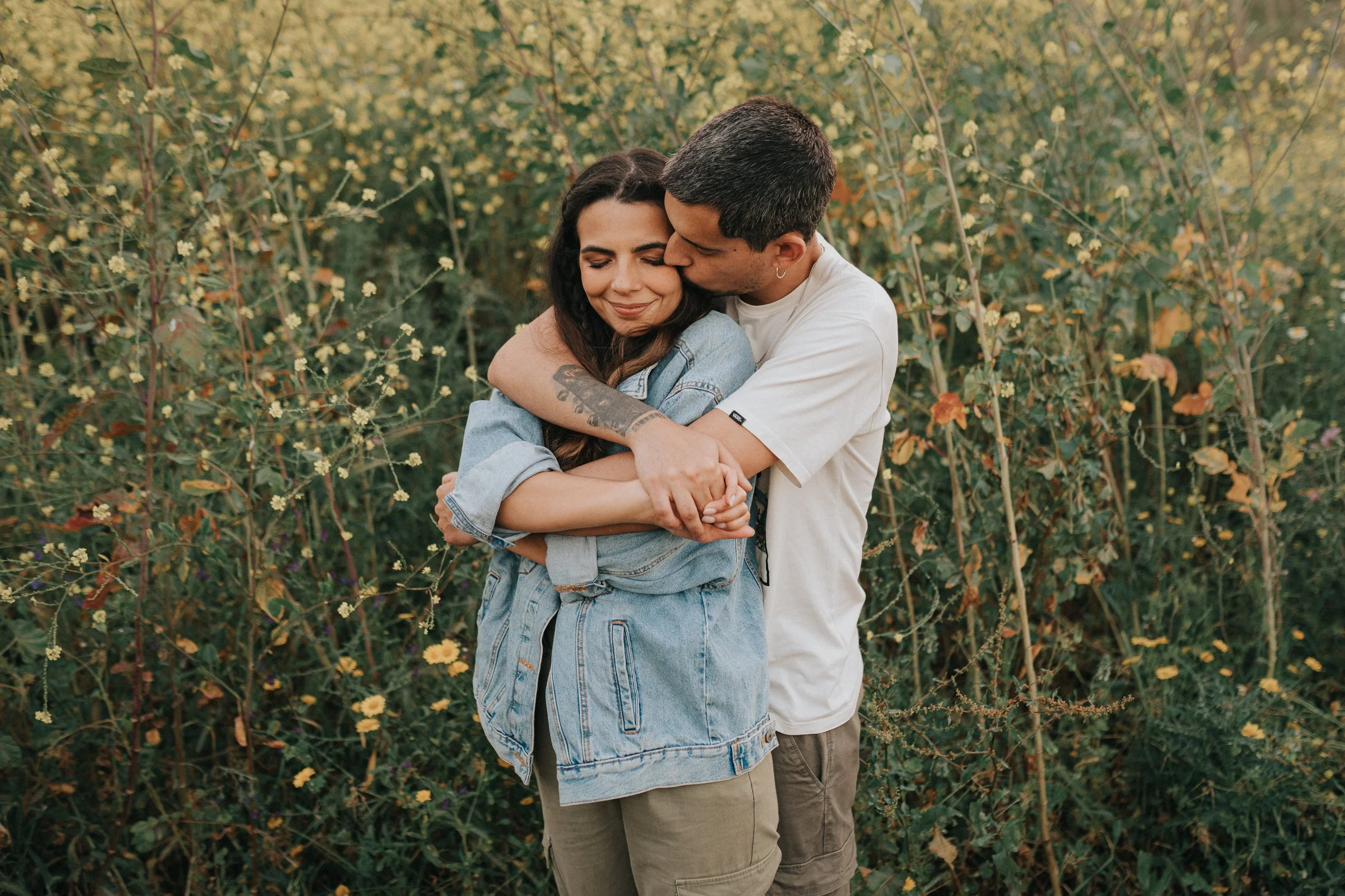 A young man and woman embracing each other outdoors in front of yellow flowering bushes.