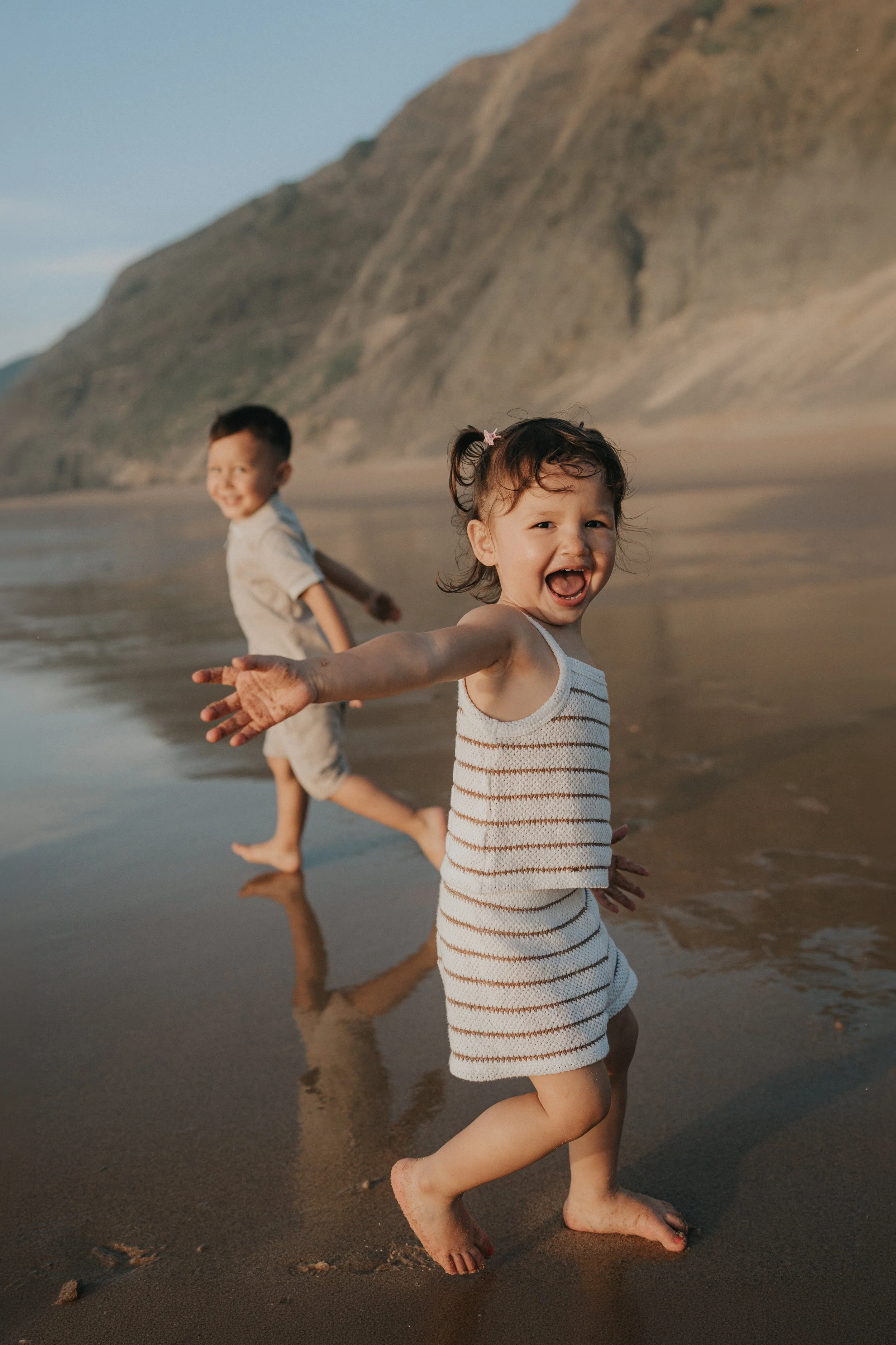 Three children running and playing on the beach near the water, with a large mountain or cliff in the background. The children are smiling and appear joyful, with the girl in the front wearing a striped dress and the other two children behind her.