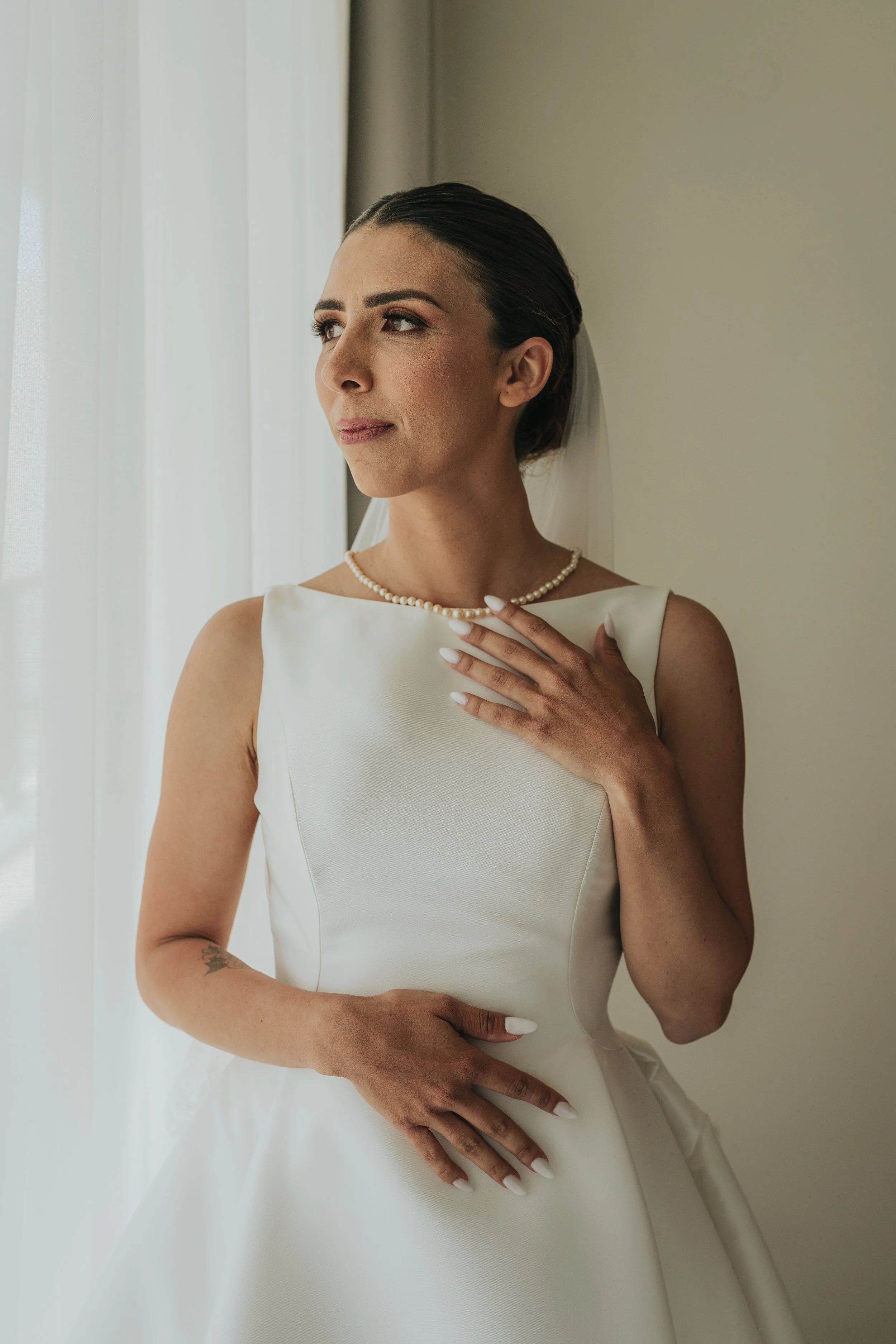 Close-up of a bride in a white wedding dress, wearing a pearl necklace, with her hand on her chest and her other hand resting on her waist, standing near a window with white curtains.