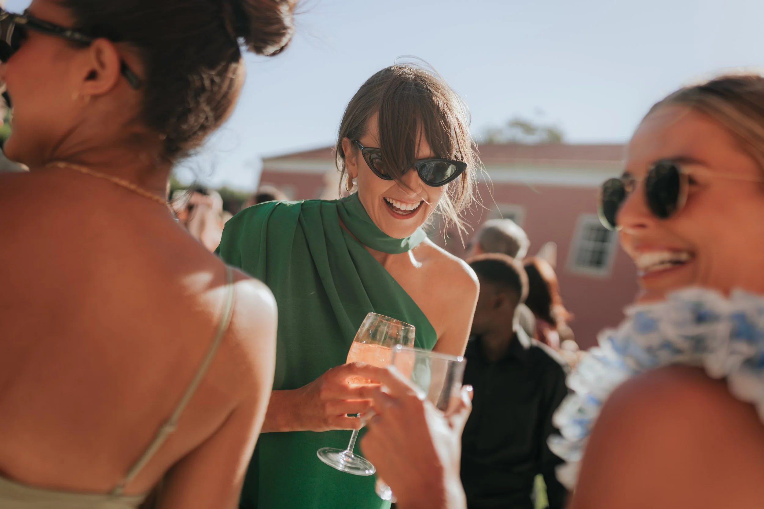 A woman in green dress holding a glass of orange-colored drink, laughing with other women at an outdoor social event.