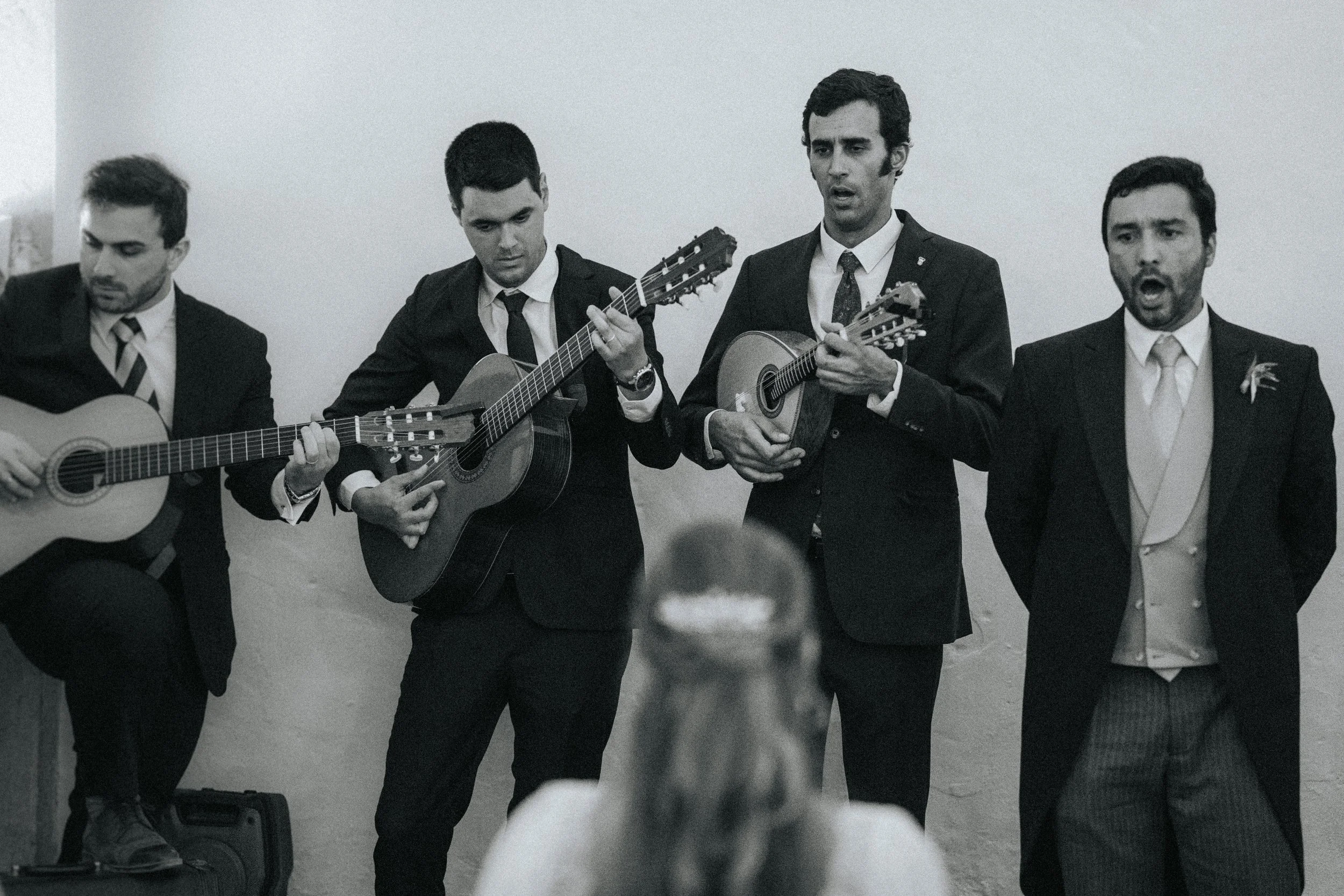 Four men in suits playing guitars and singing at a performance, standing in front of a plain, light-colored wall.