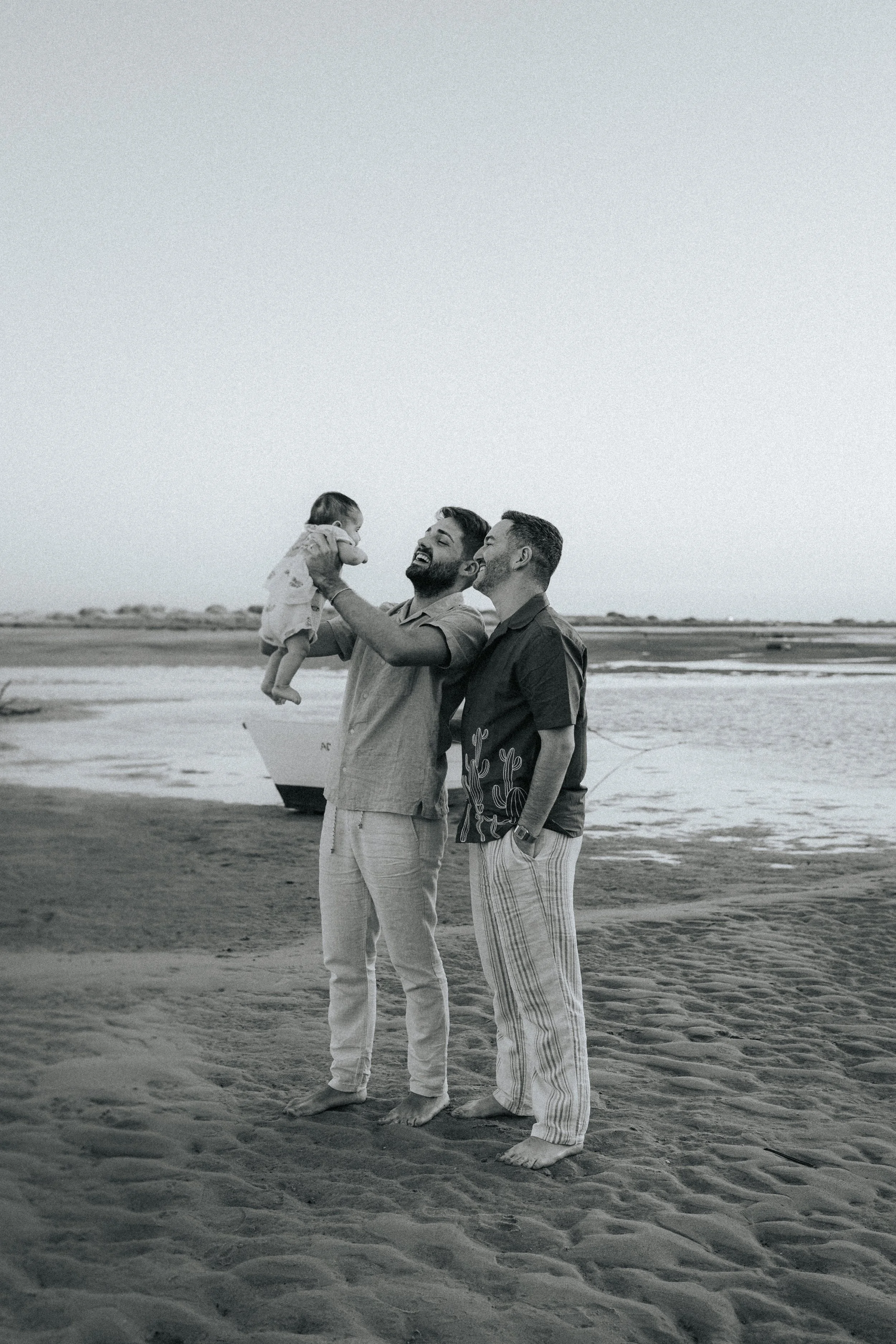 Two men standing barefoot on the sand at the beach, one holding a child up in the air, with the ocean and a boat in the background, black and white photo.