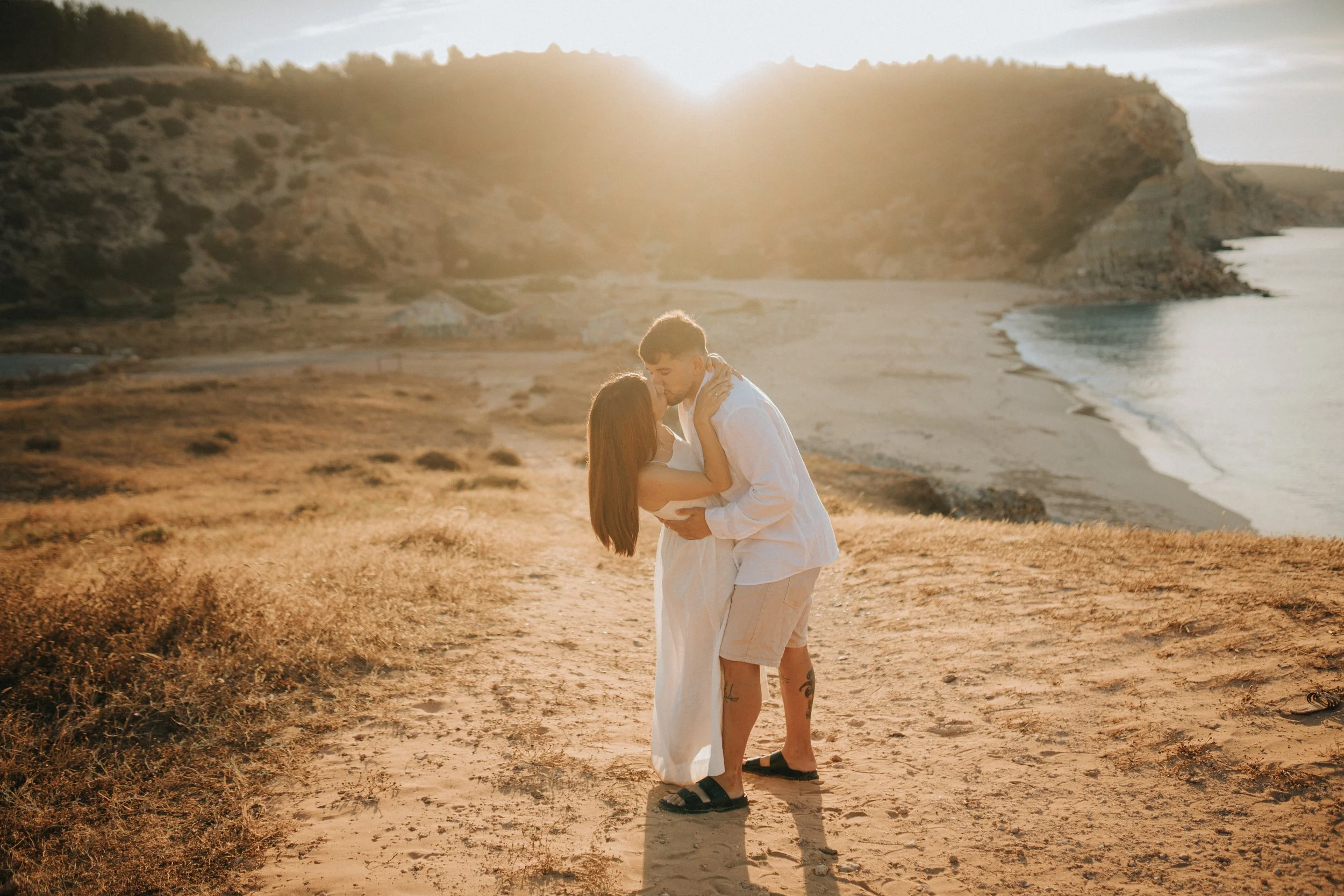 A couple is kissing and embracing on a sandy beach during sunset. The woman has long red hair, and the man has short brown hair. They are dressed in white clothing, with the woman in a dress and the man in a shirt and shorts. The landscape features c