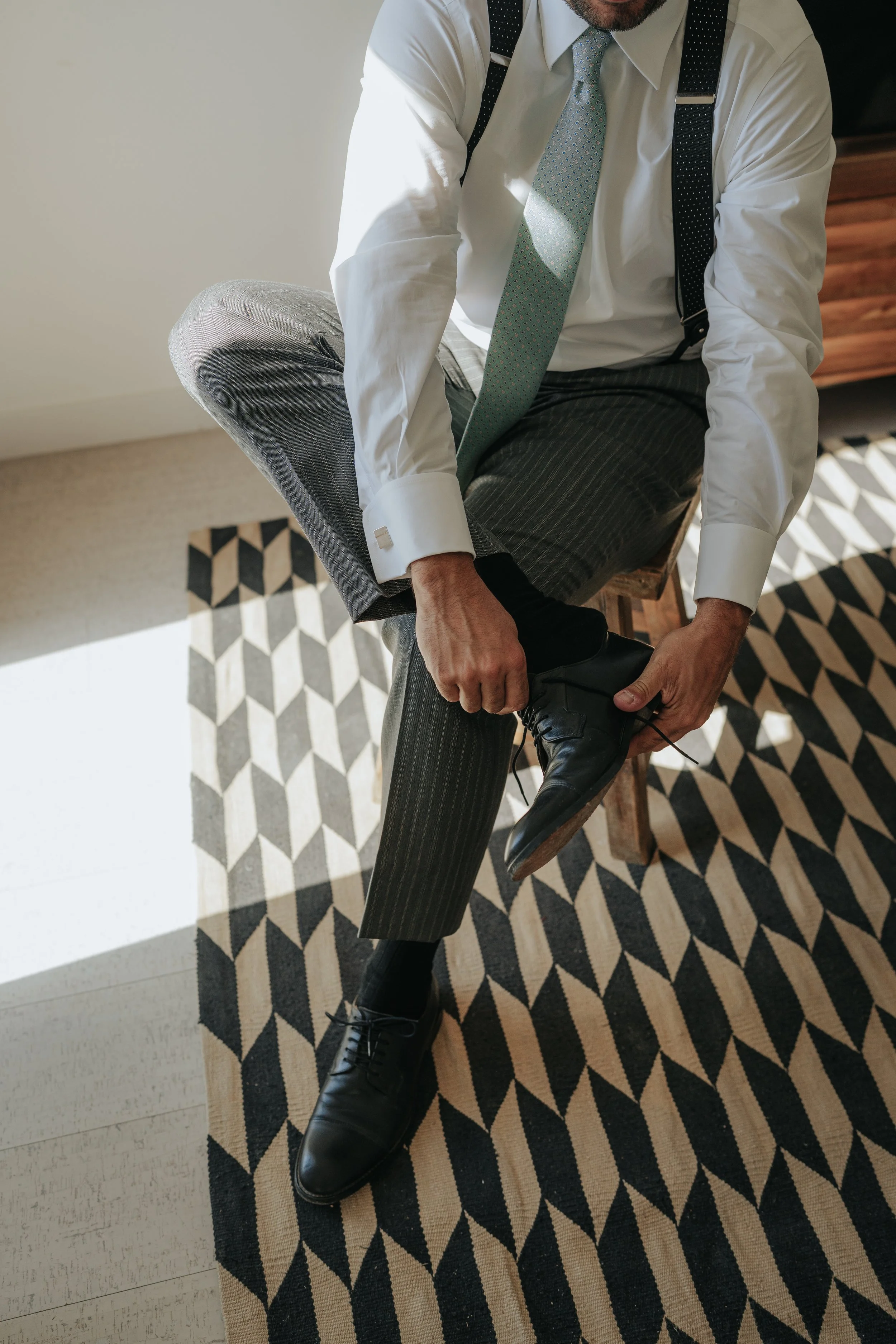 A man in a white dress shirt, gray striped pants, and a light blue patterned tie is sitting on a wooden stool, tying his black dress shoes. The man is also wearing black suspenders, and his other leg is crossed over his knee. He is positioned on a pa