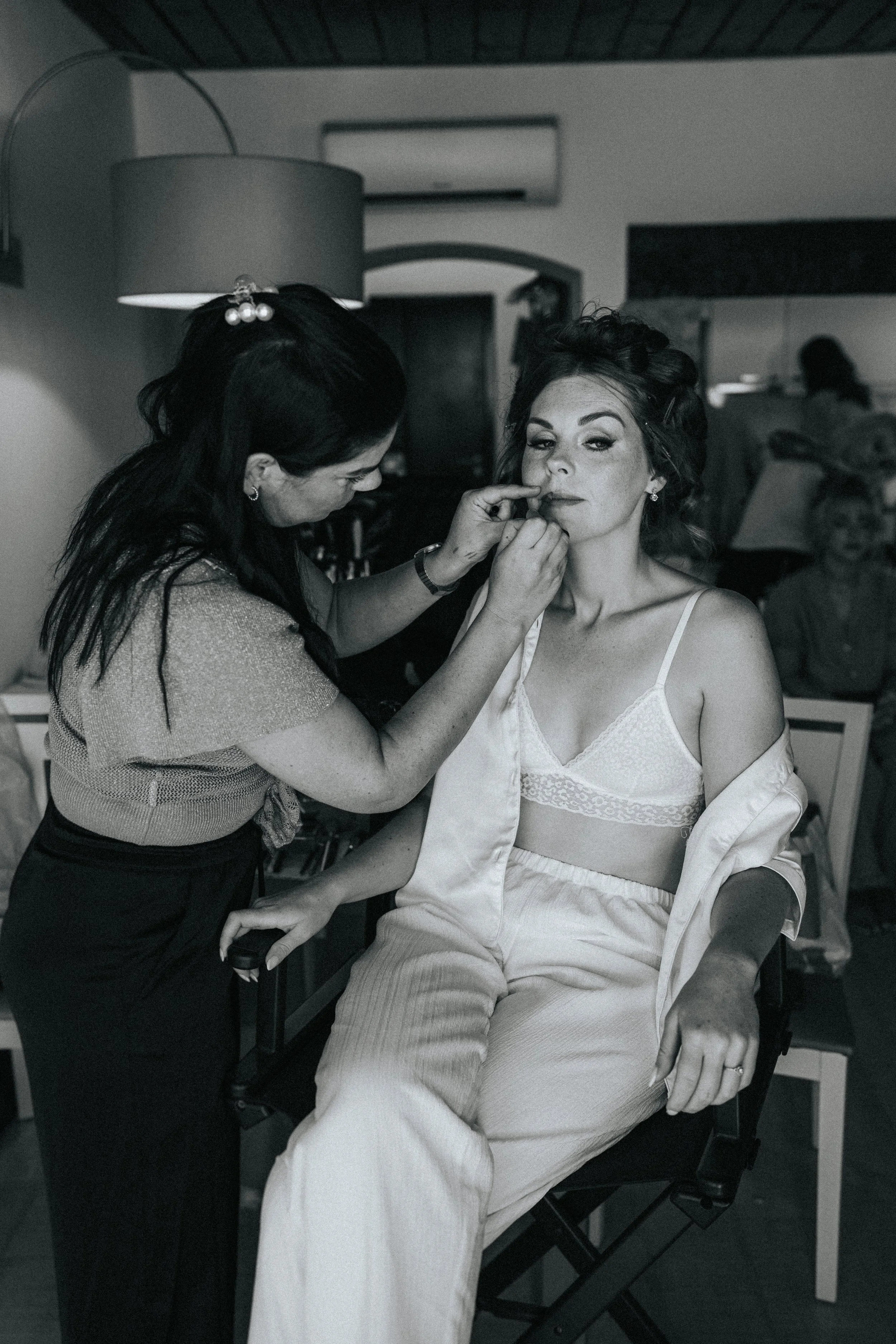 A woman sitting in a chair, getting her makeup done by a makeup artist in a room with a large lamp, a television, and a doorway in the background.