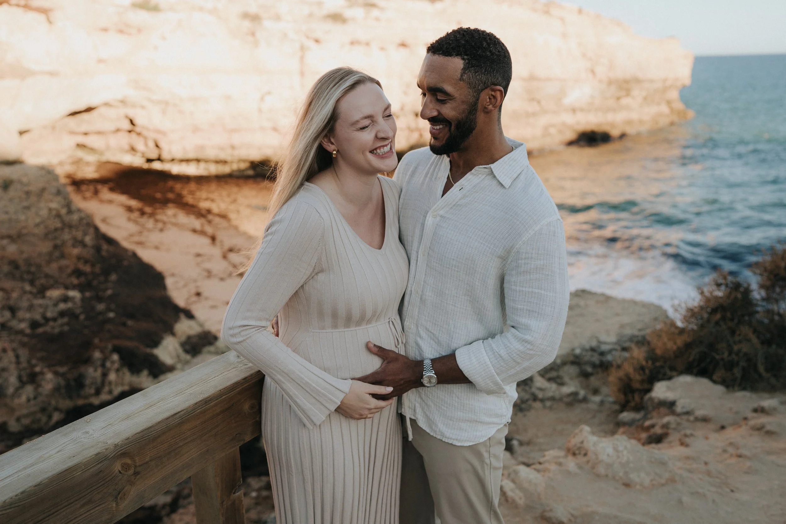 A happy pregnant woman and a smiling man embracing near the ocean on a sunny day, standing on a wooden walkway.
