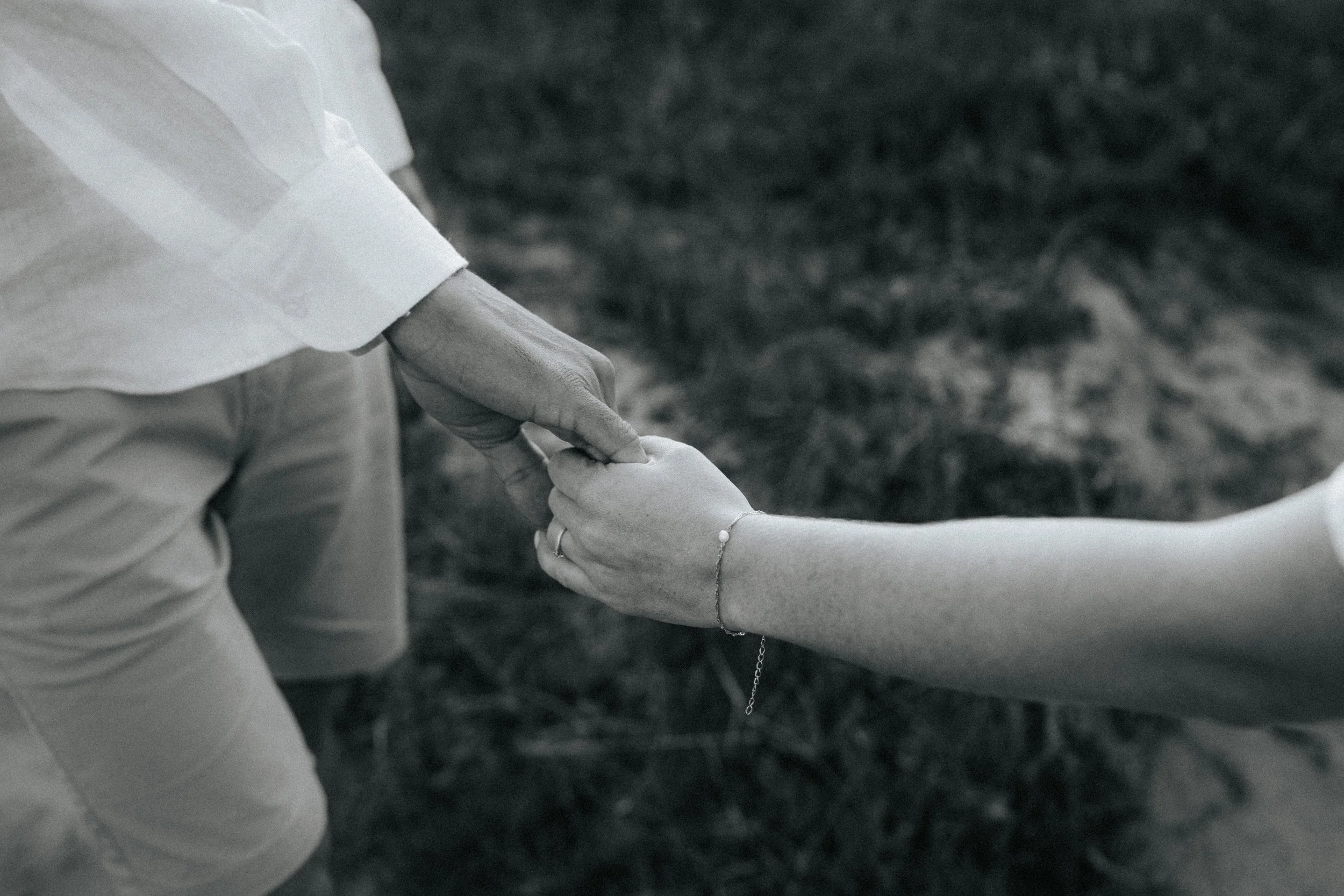 A couple holding hands outdoors, with focus on their joined hands and forearms, one wears a bracelet and the other has a ring.
