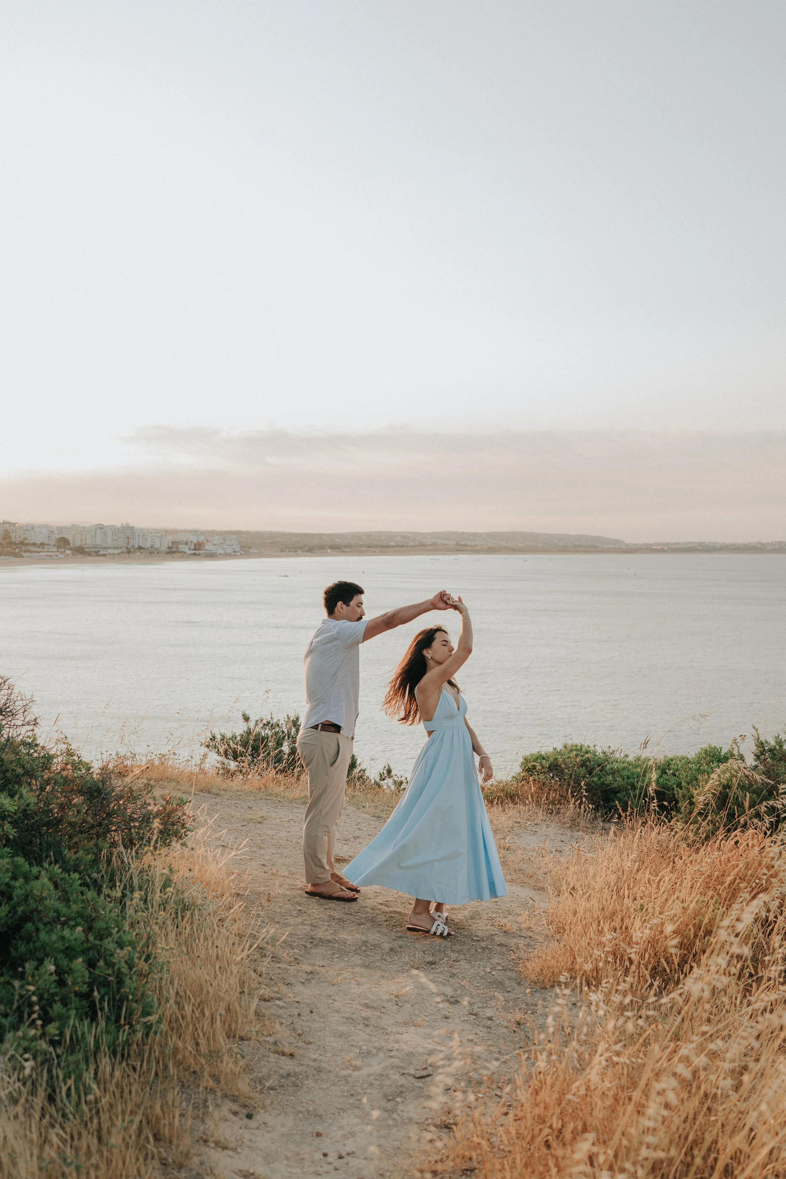 A couple dancing outdoors near a body of water during sunset, with the backdrop of a city skyline.