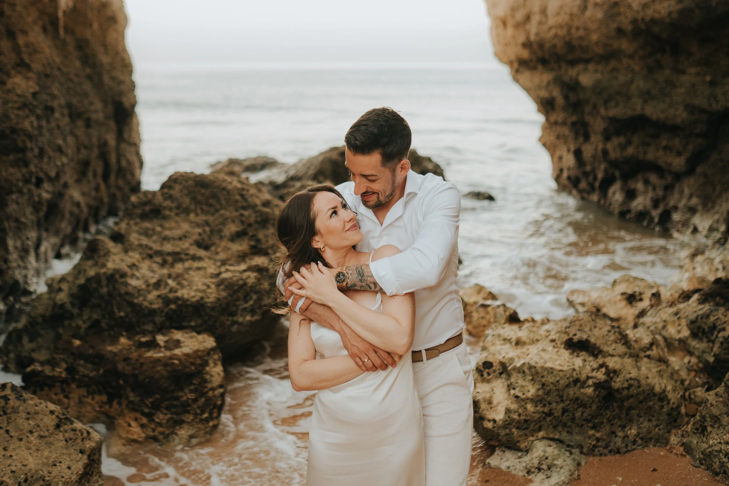 A couple embracing on a rocky beach, with the ocean in the background, framed by large rock formations.