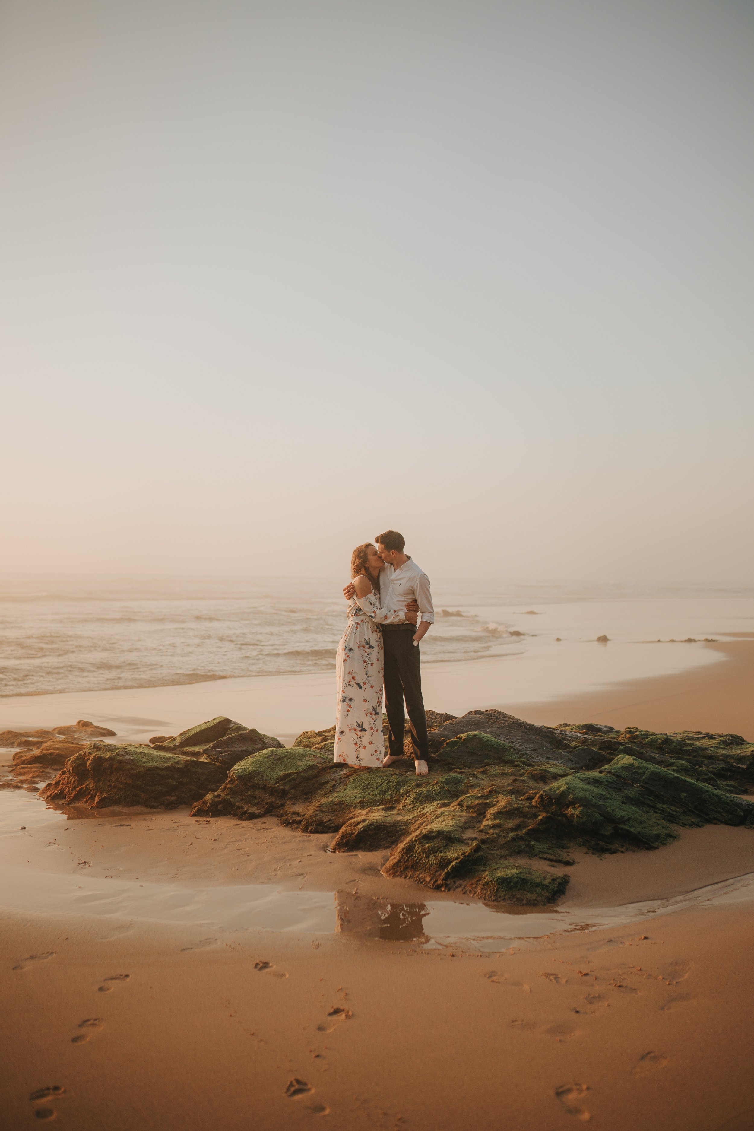 A couple embracing on rocks at the beach during sunset, with the ocean in the background and footprints in the sand in the foreground.