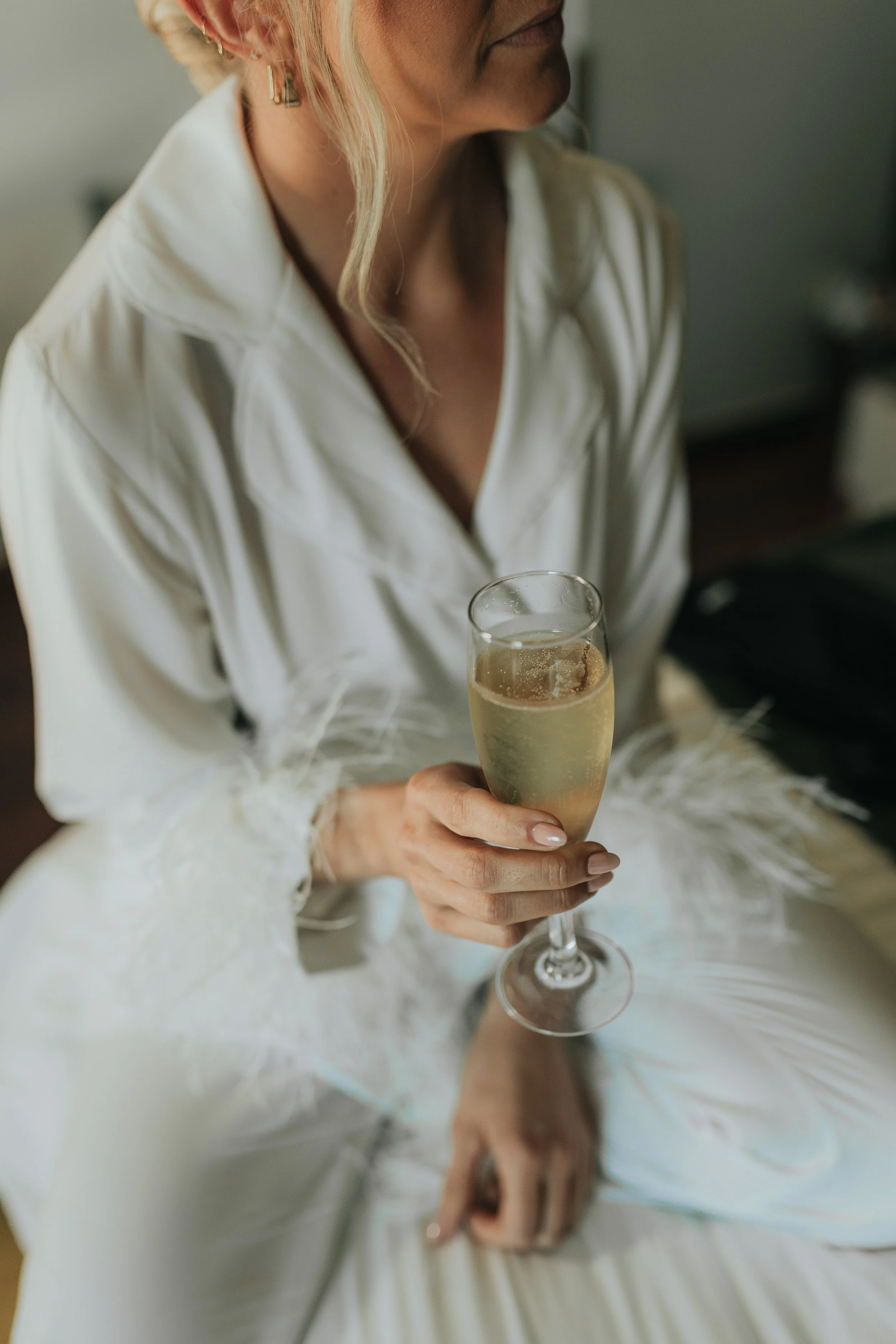 A woman in a white dress holding a glass of champagne, sitting on a bed.