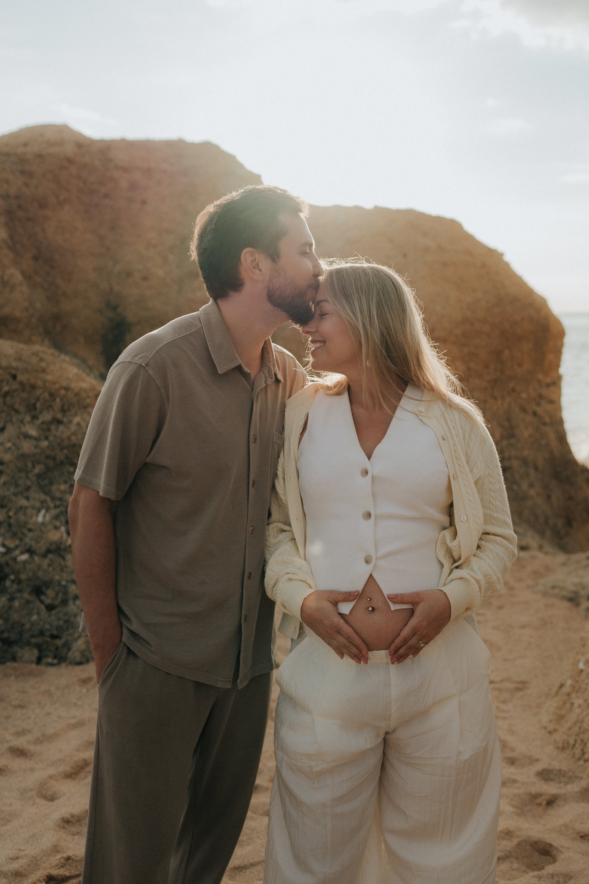 A man and a woman standing close together on a beach with large rocks in the background. The man is kissing the woman's forehead, and the woman is smiling with her hands on her pregnant belly.