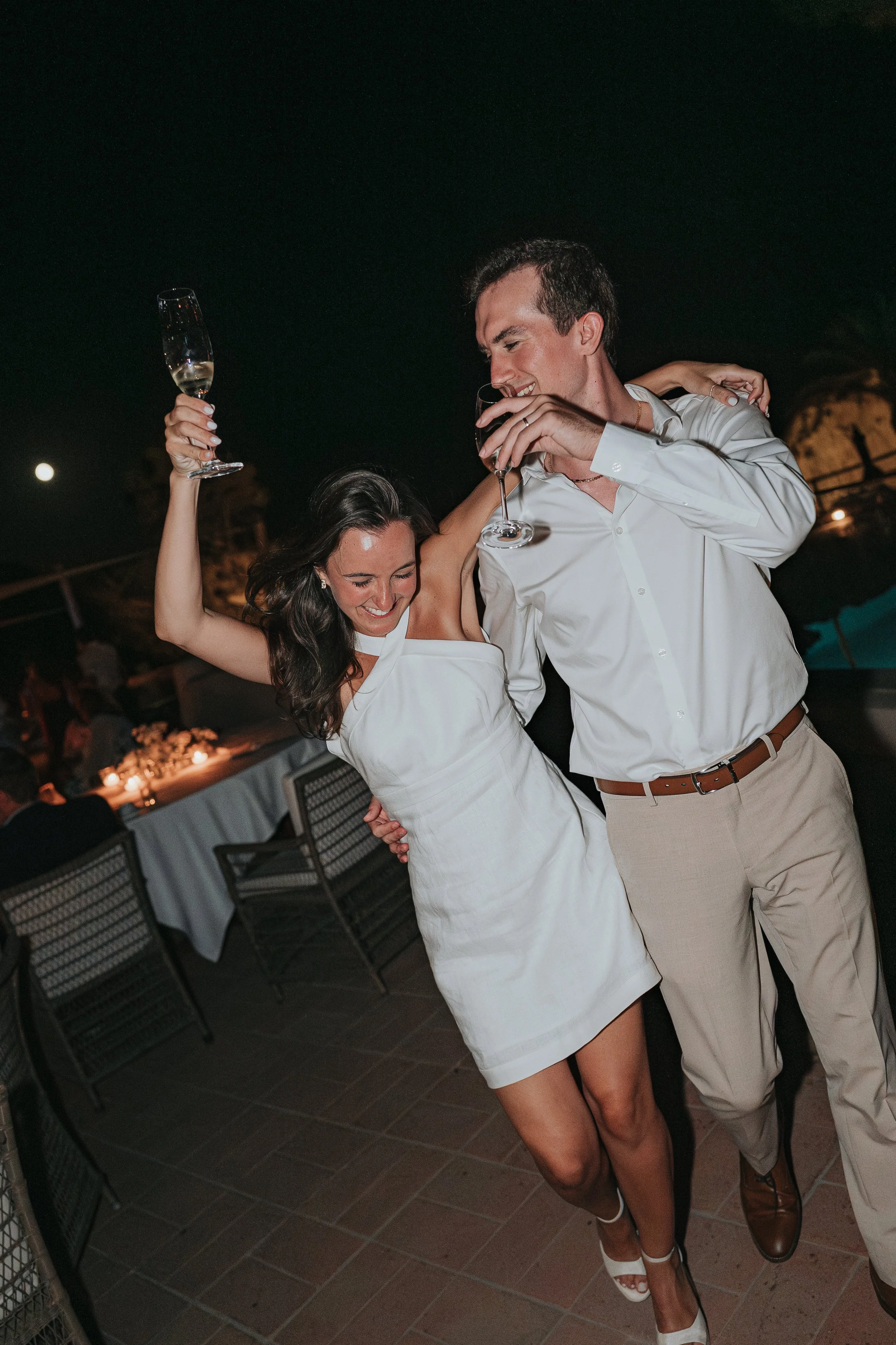 A man and woman celebrating at night, smiling and holding glasses of champagne, with the woman arm around the man's shoulder and her other arm raised holding a glass, in an outdoor setting with chairs and table in the background.